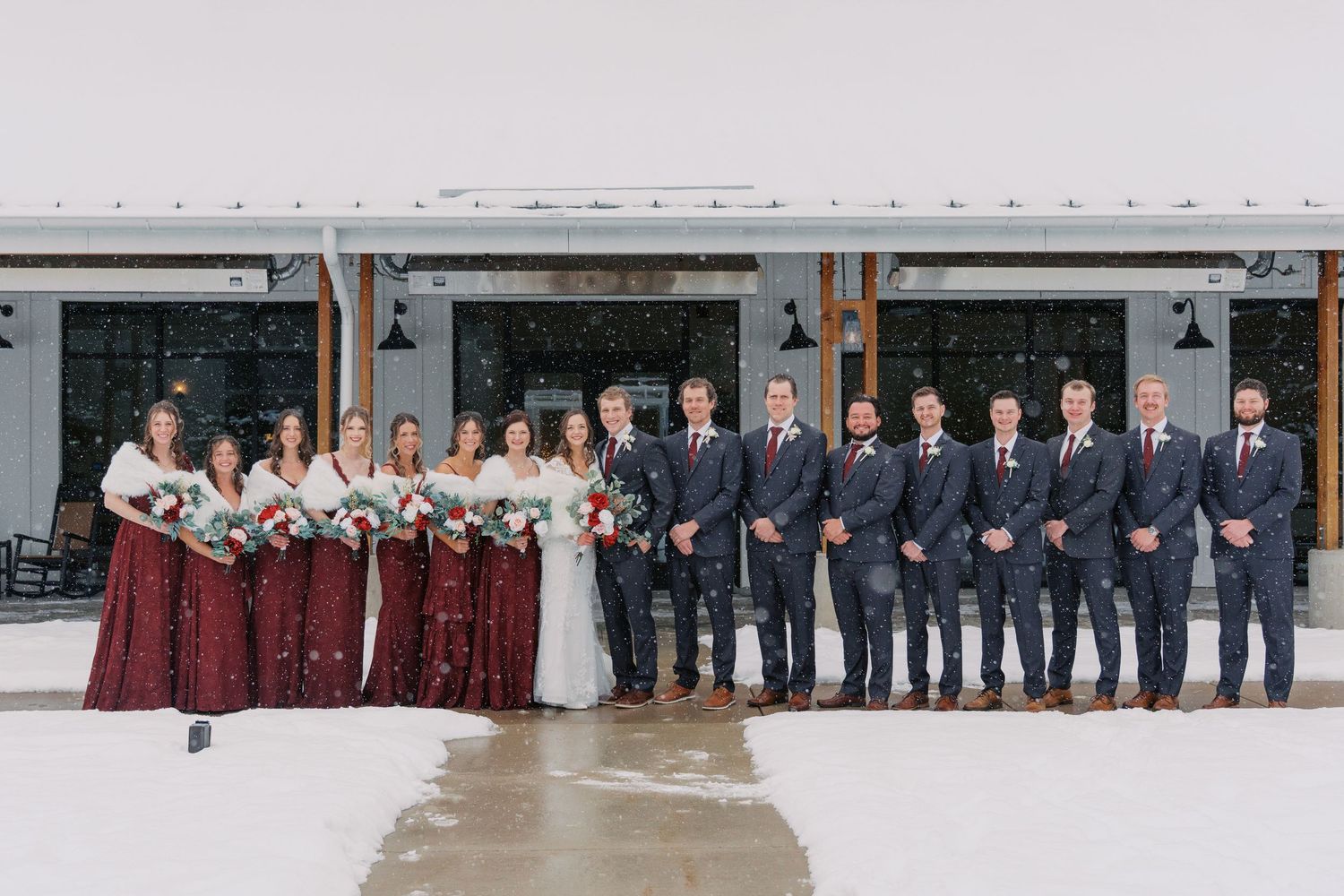 Wedding party lined up outdoors in burgundy and navy attire against grey building.