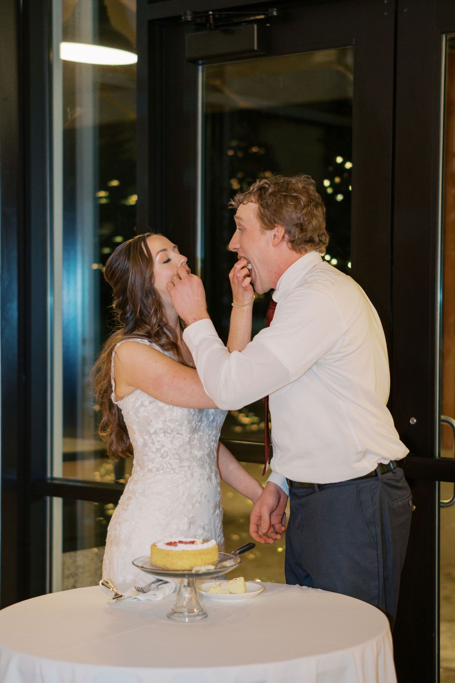 Newlyweds share cake at wedding reception table with festive lights in background.