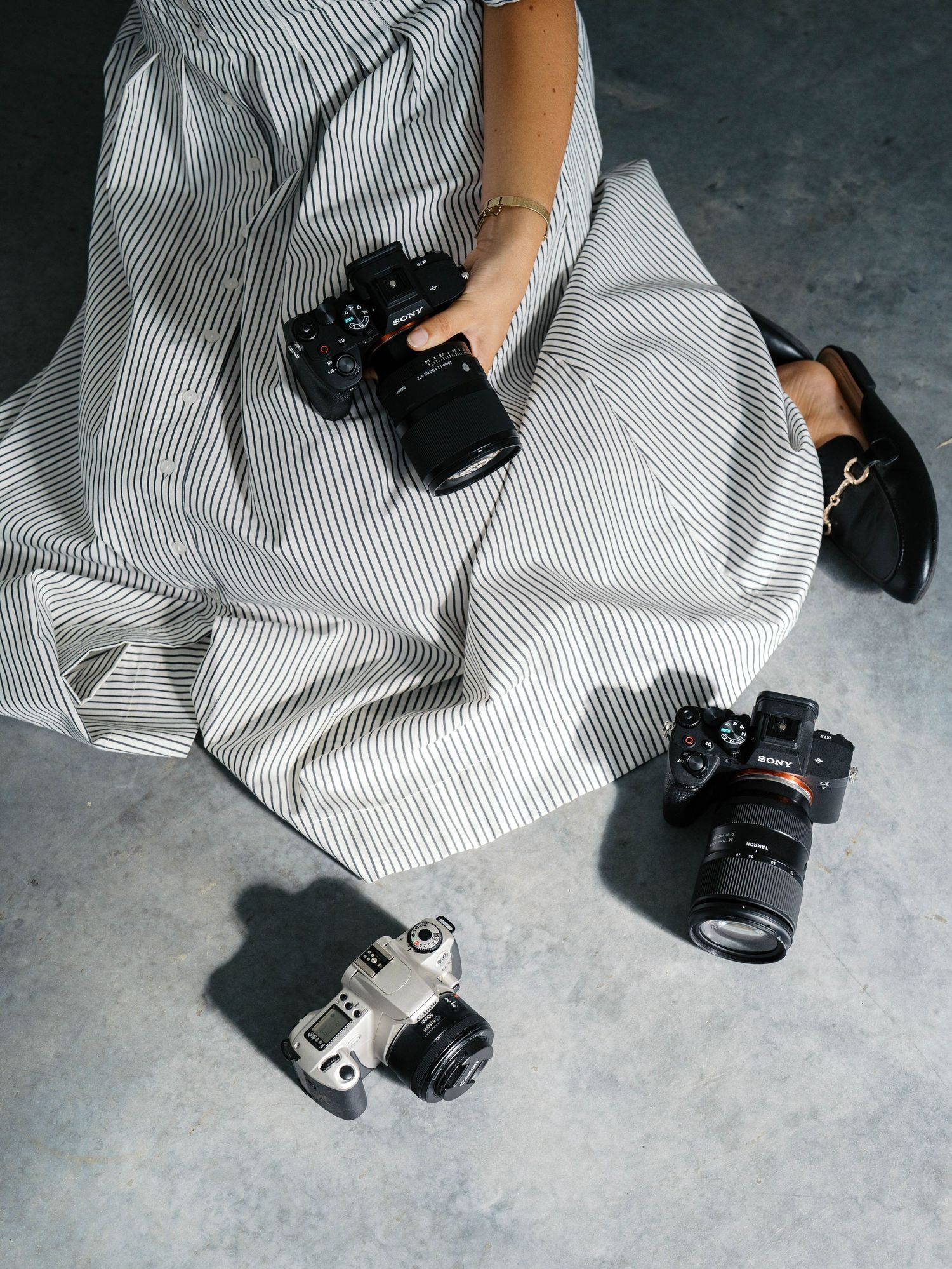 Woman in striped dress sitting on floor holding DSLR camera with two additional cameras placed nearby.