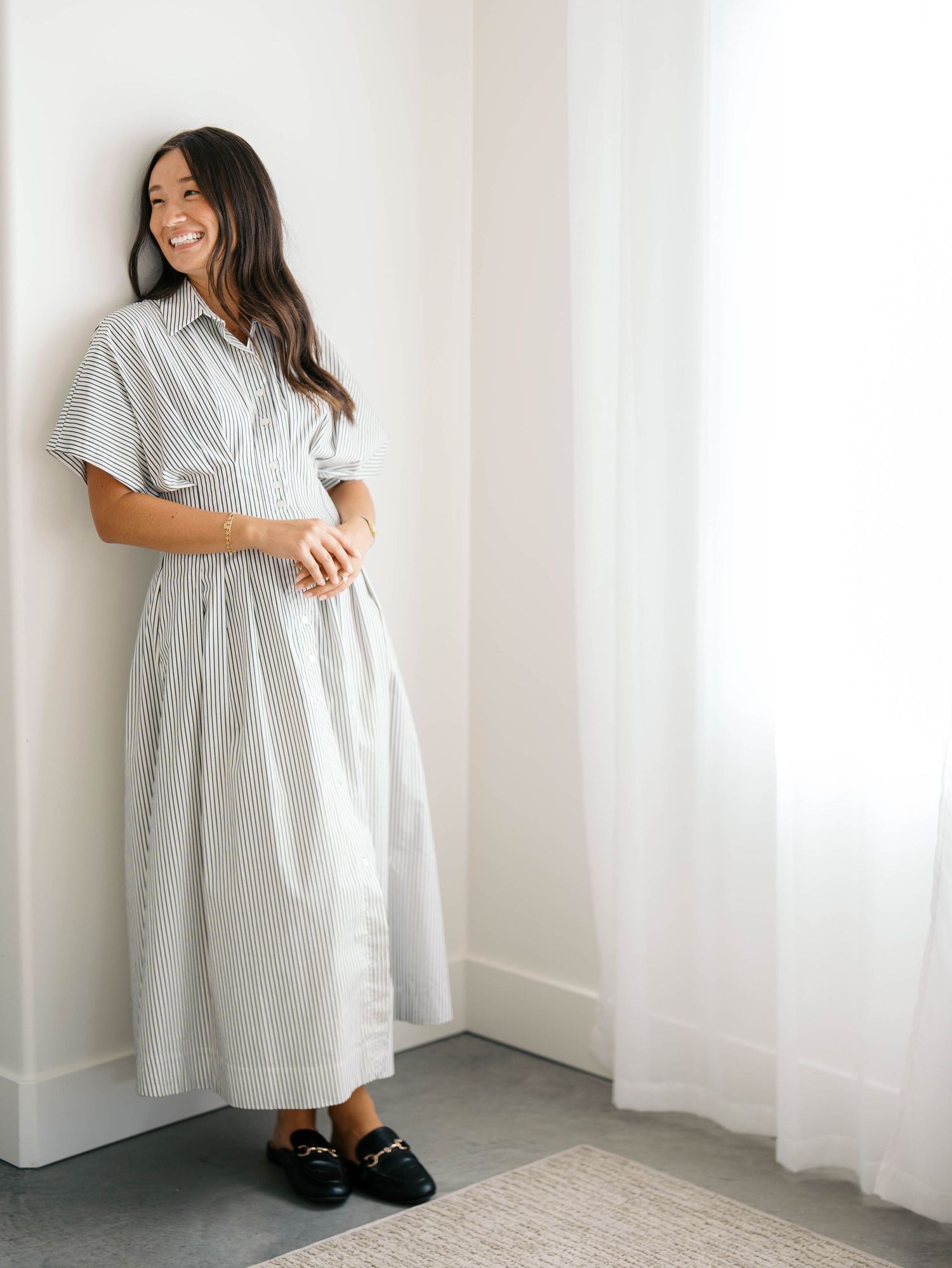 Woman smiling in a striped midi shirtdress with short sleeves, paired with black loafers, leaning against a white wall.