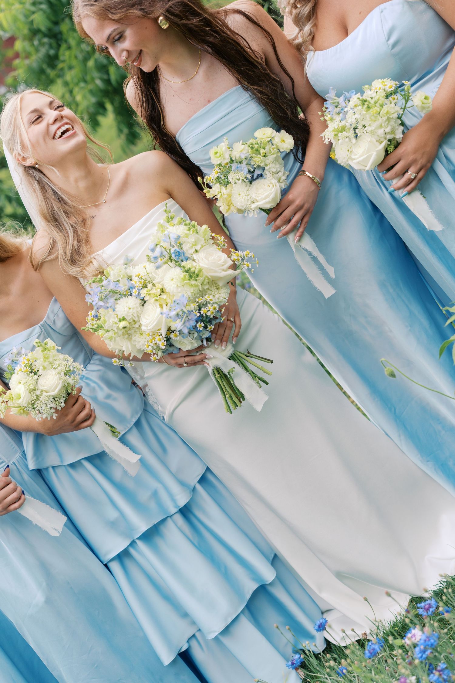 Bridesmaids in light blue strapless gowns holding white and blue floral bouquets, laughing outdoors at a wedding.