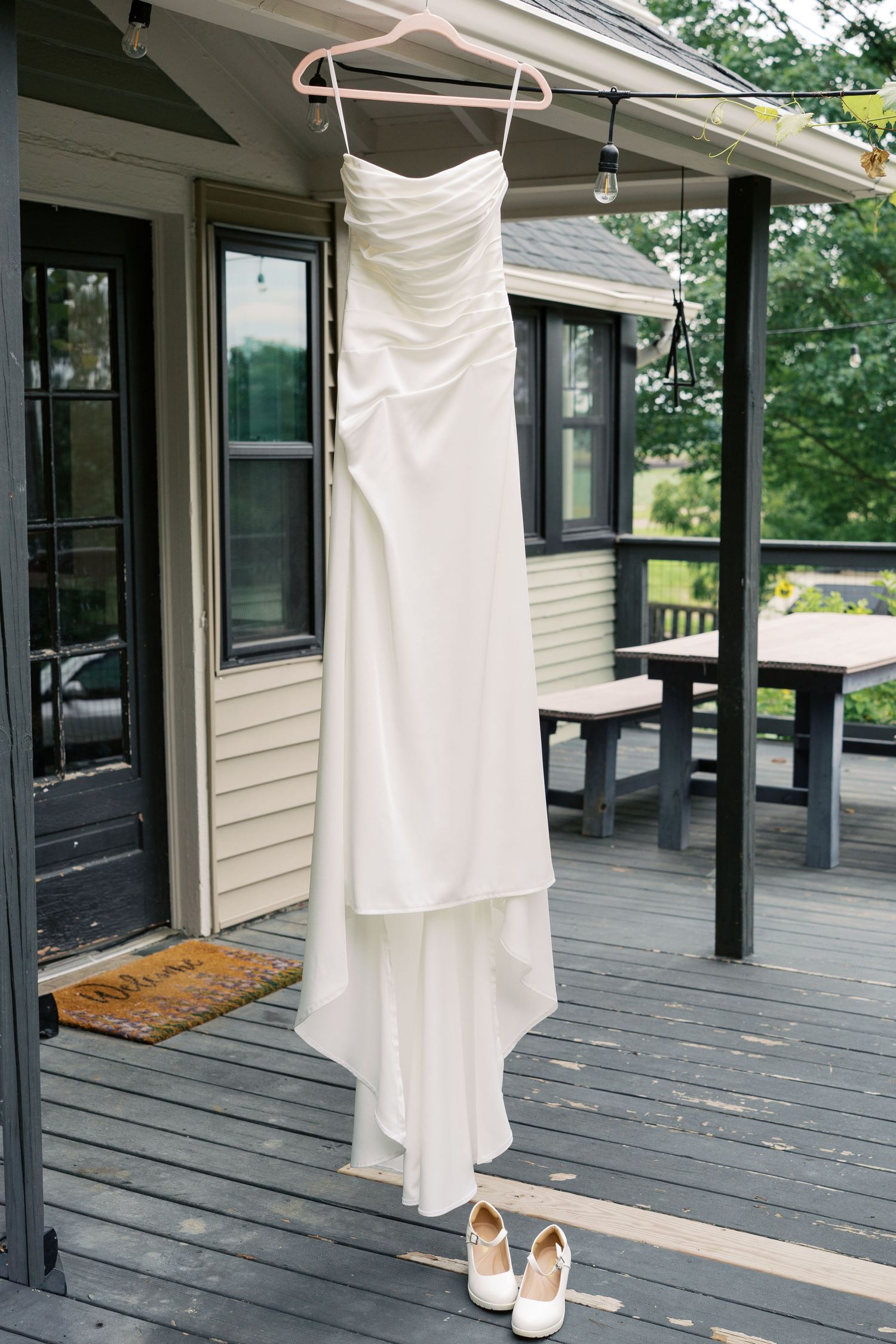 White wedding dress hanging on a wooden porch with white bridal heels below on a rustic outdoor deck.