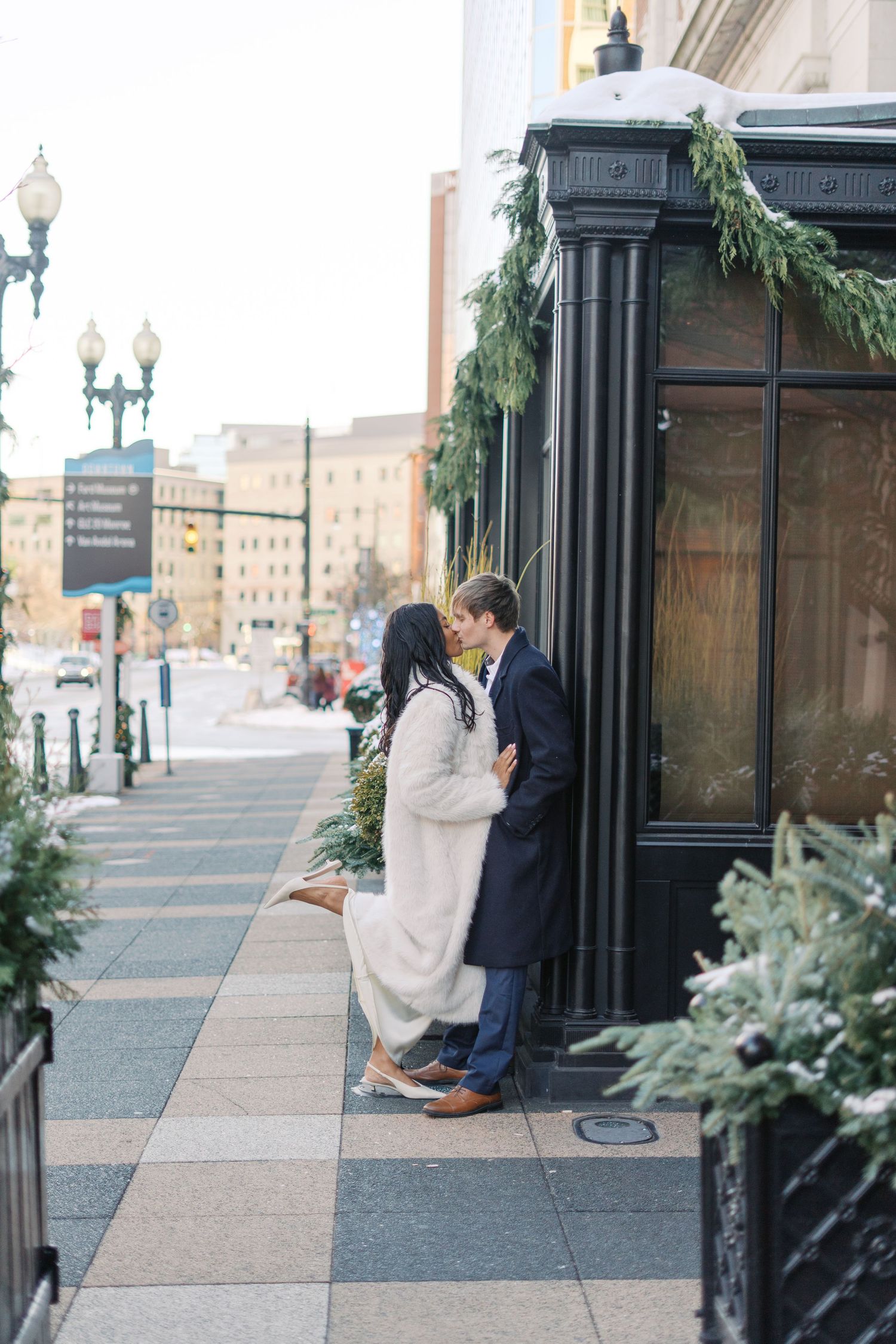 A couple shares a tender moment beside an ornate black pillar on a city sidewalk.