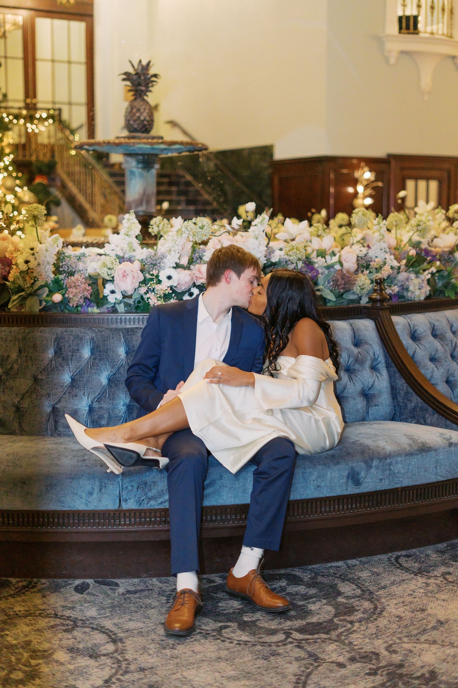 Couple lounging on vintage blue velvet couch with floral arrangements.