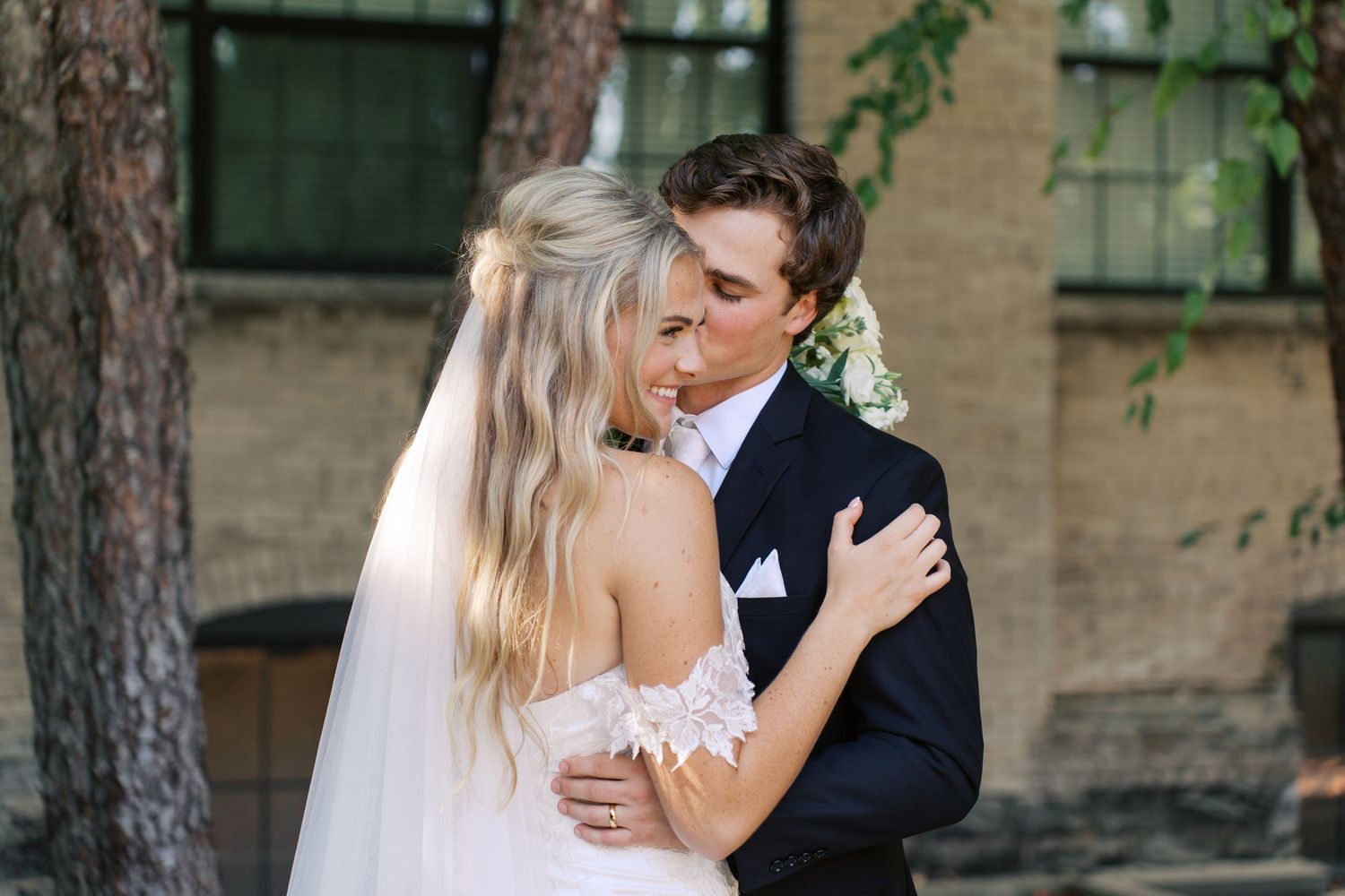 Romantic outdoor wedding portrait with couple embracing against brick building backdrop.