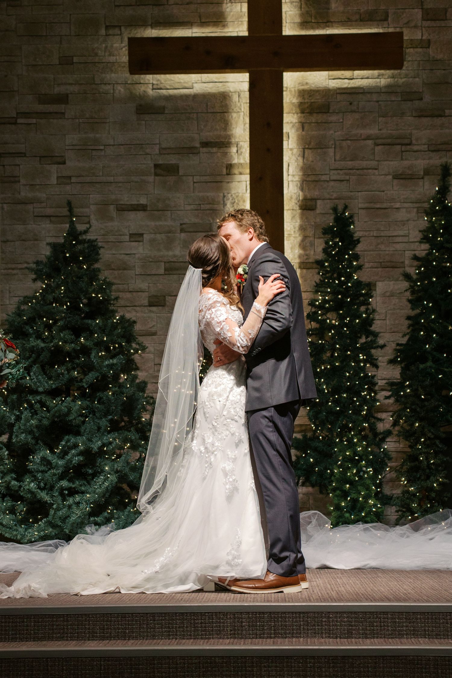 A romantic wedding moment captured in front of a brick wall with Christmas trees and decorative lighting.