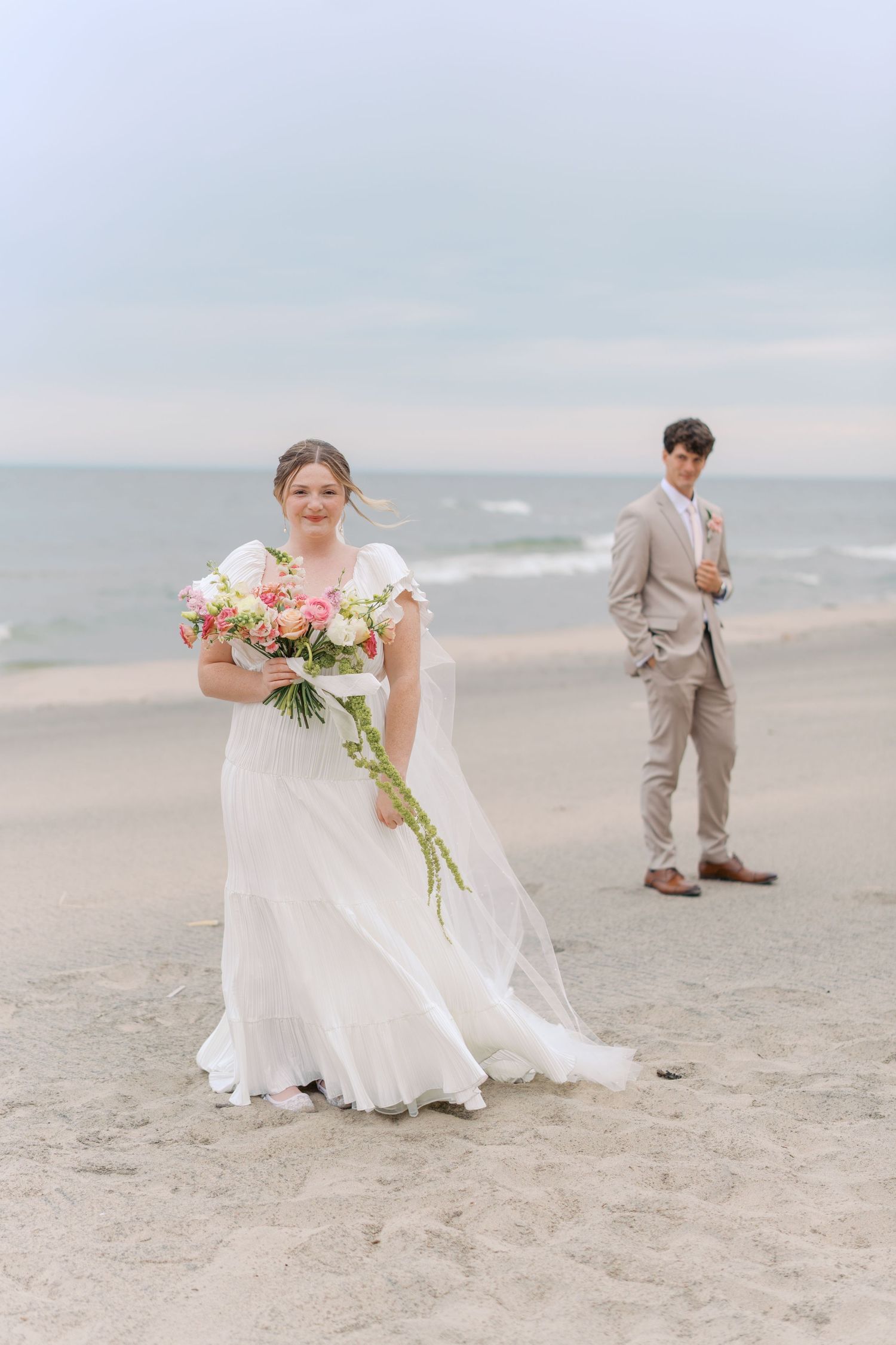 Bride in white gown holds pink bouquet on beach, groom in tan suit stands behind near ocean.