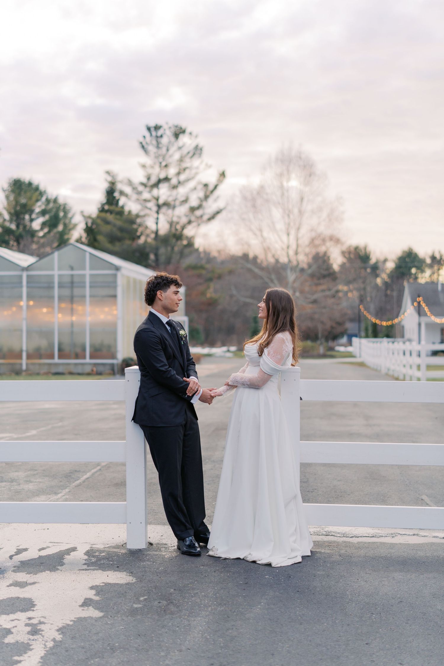 A bride and groom hold hands facing each other in front of a white fence at an outdoor wedding venue.