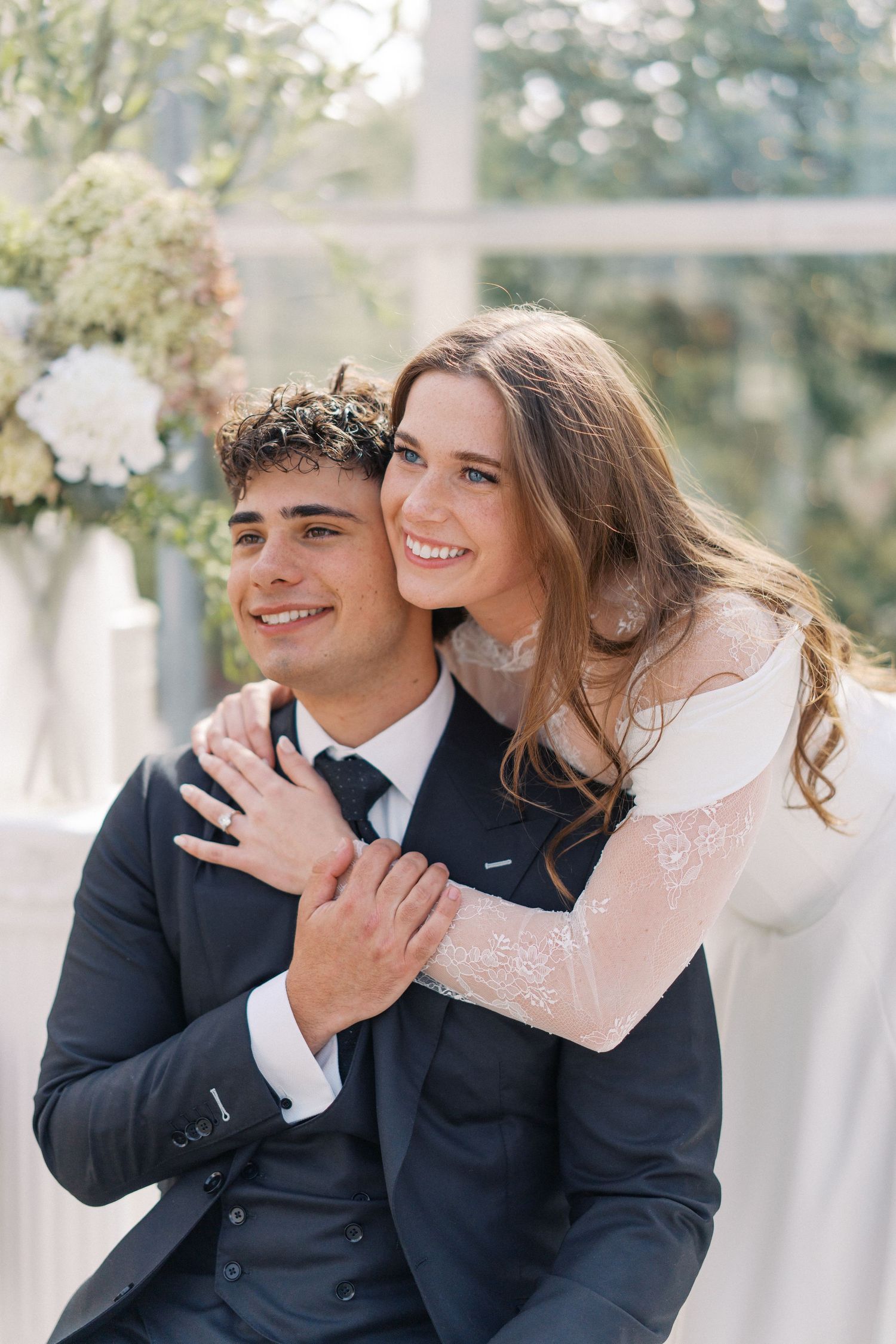 A smiling bride in a lace gown embraces a groom in a navy suit at an elegant floral wedding ceremony.
