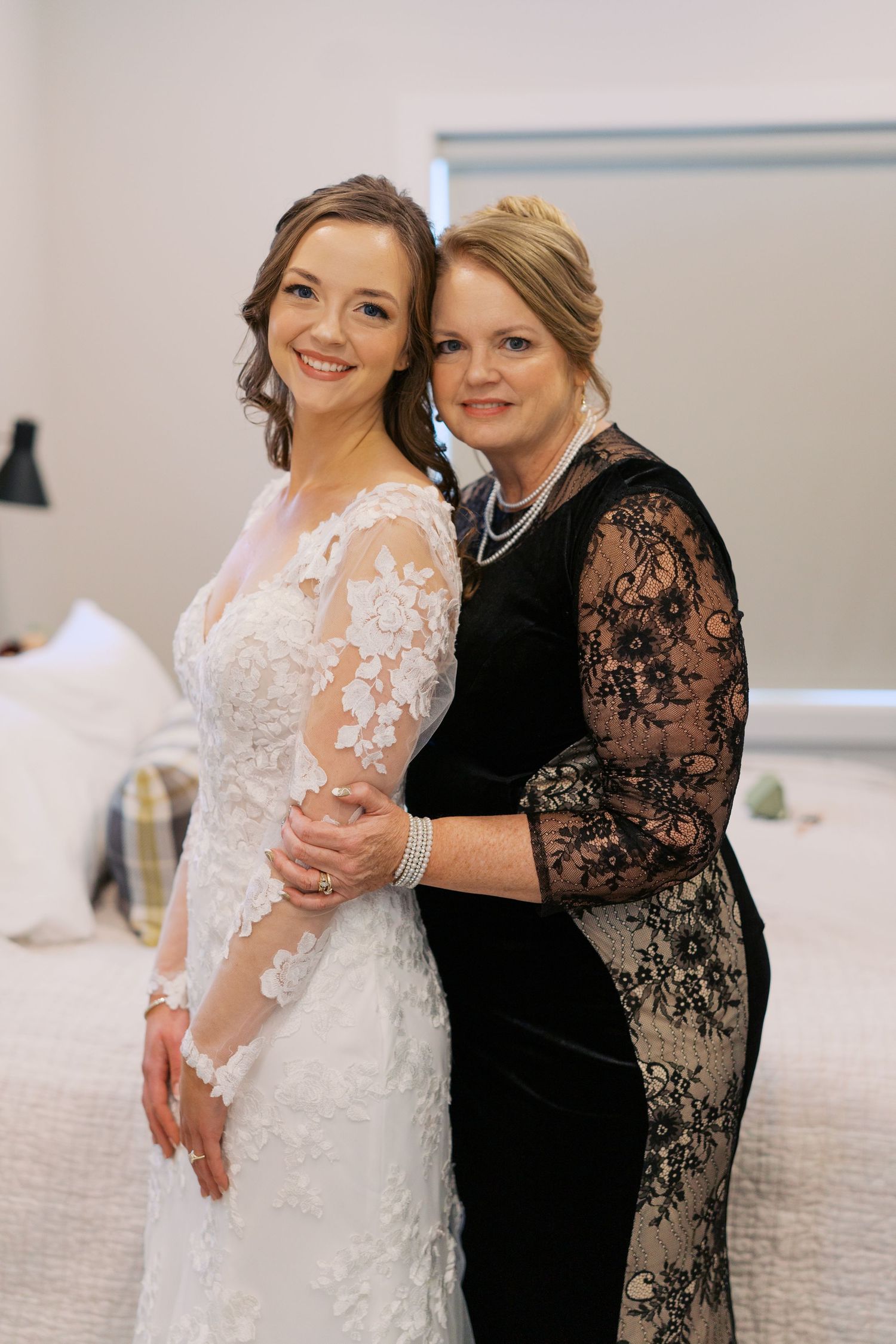 Emotional moment between bride in lace gown and woman in black lace dress during wedding preparations.