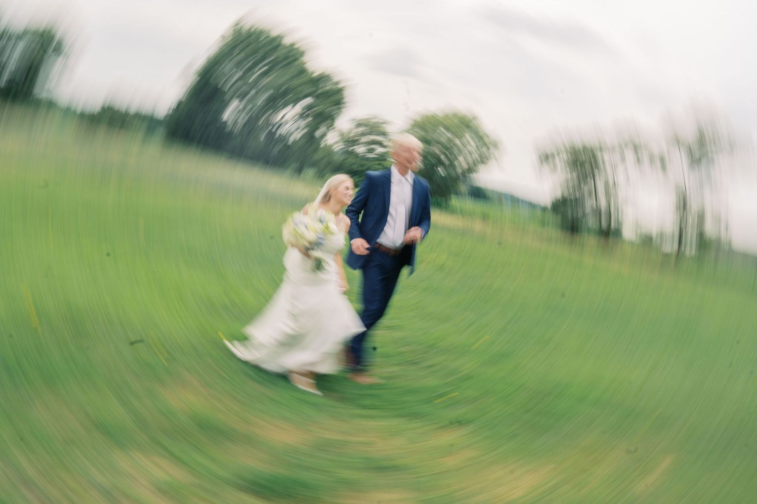 Bride and groom running joyfully through a lush green field on their wedding day, captured with a dreamy spin blur effect.