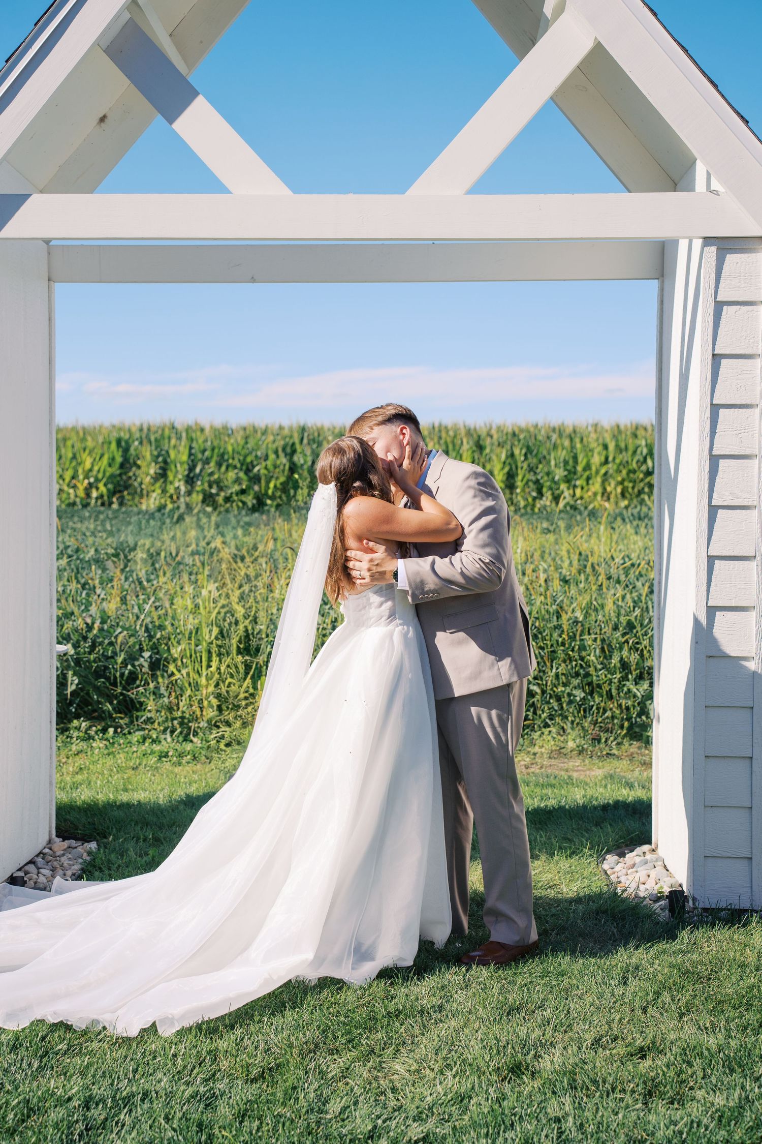 Wedding couple poses under a white pergola with cornfields in the background.