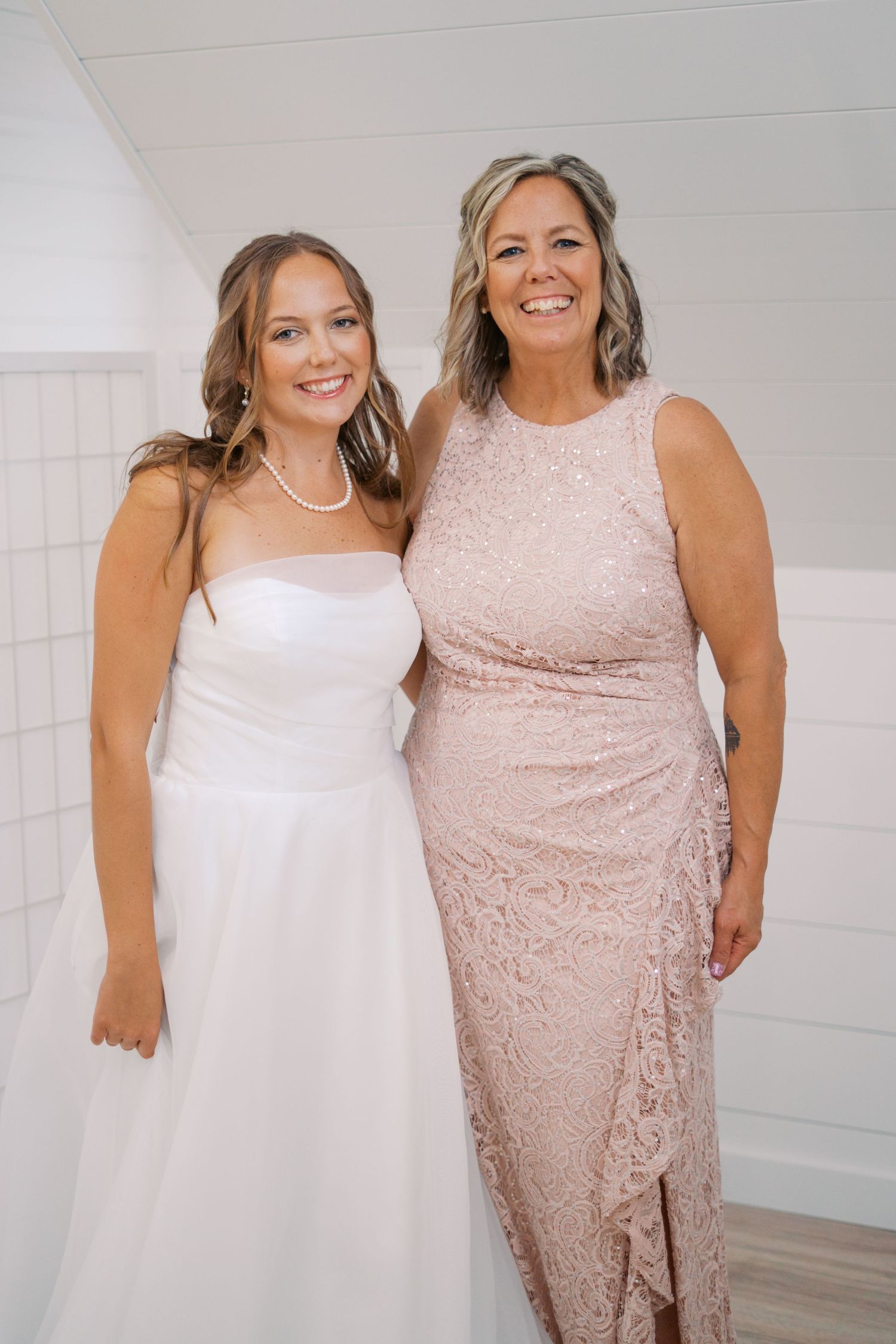 Two women pose together wearing formal dresses in blush pink and white against a light background.