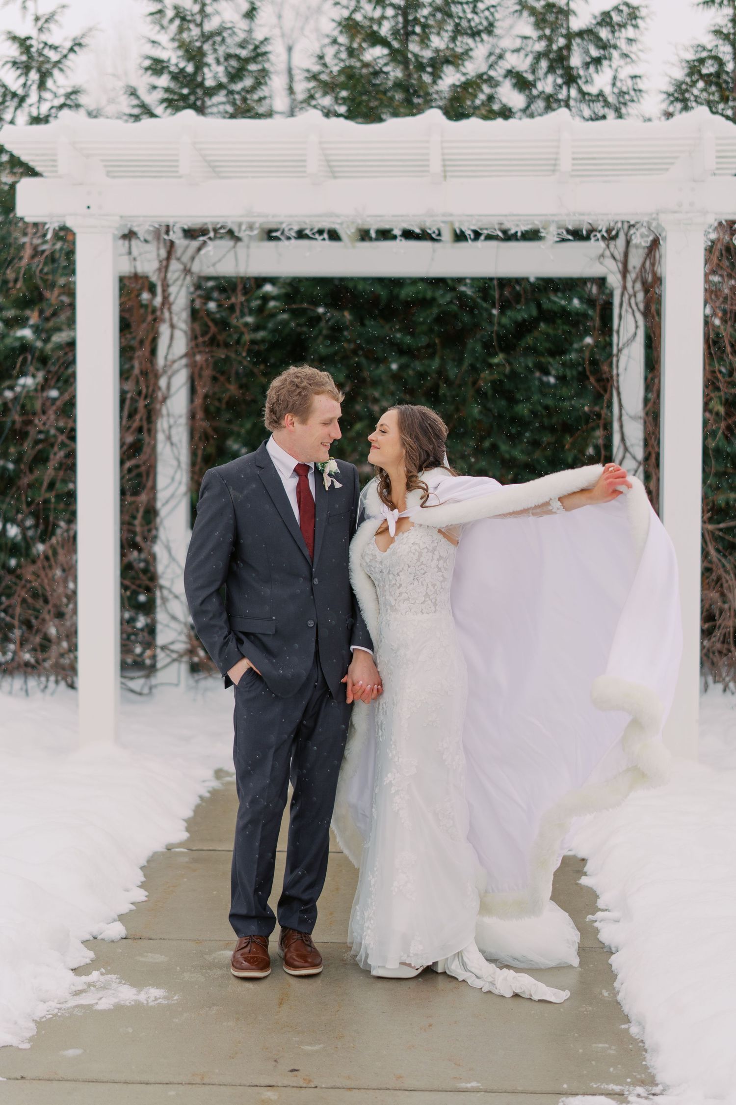 Bride playfully twirls her dress next to groom under white pergola in snowy setting.