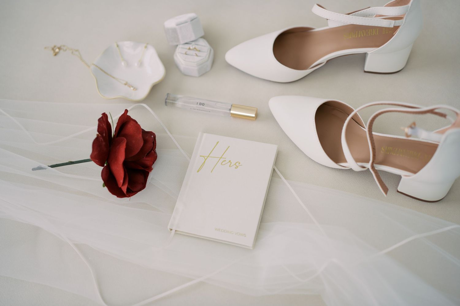 Flat lay of white wedding accessories including shoes and stationery on light surface.
