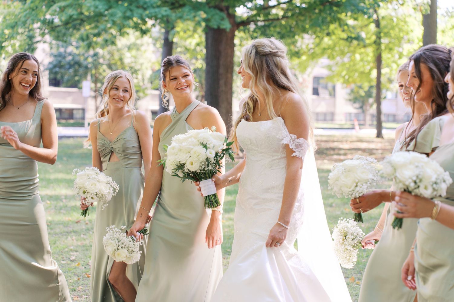 Bridesmaids in sage dresses gather around bride for casual outdoor wedding photos.