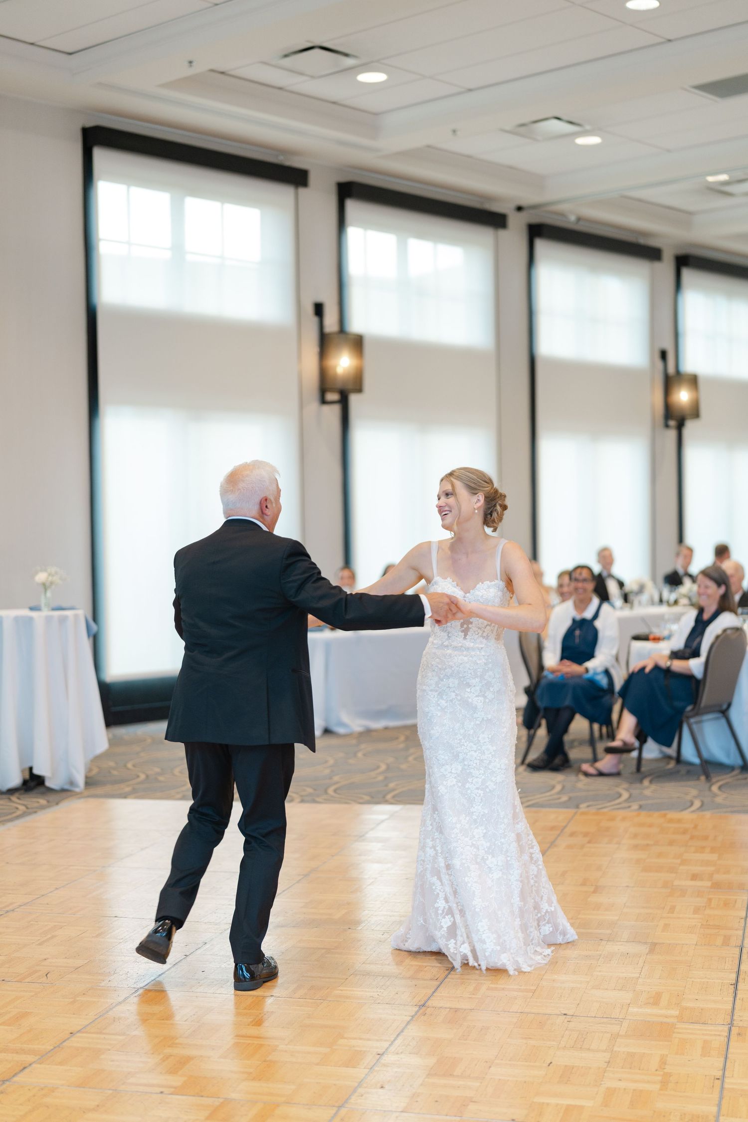 A wedding couple dances together on a wooden floor in an elegant reception hall with white decor.