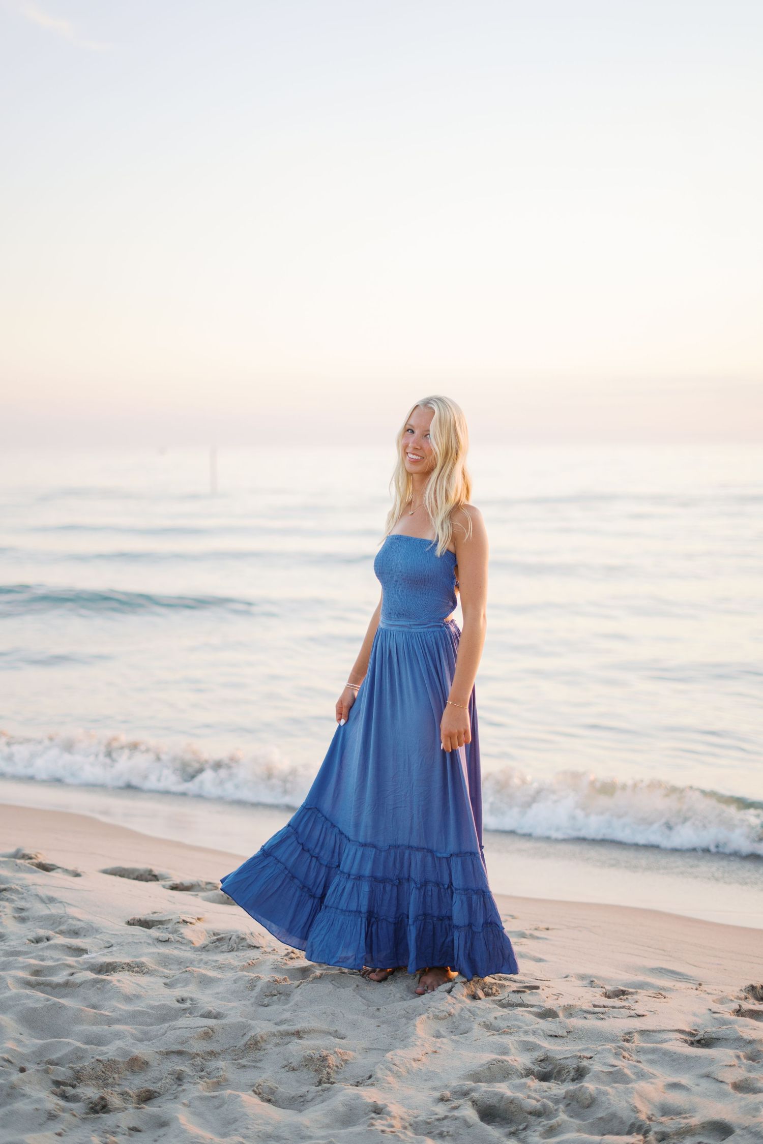 Woman in flowing blue dress twirls on sandy beach at sunset.