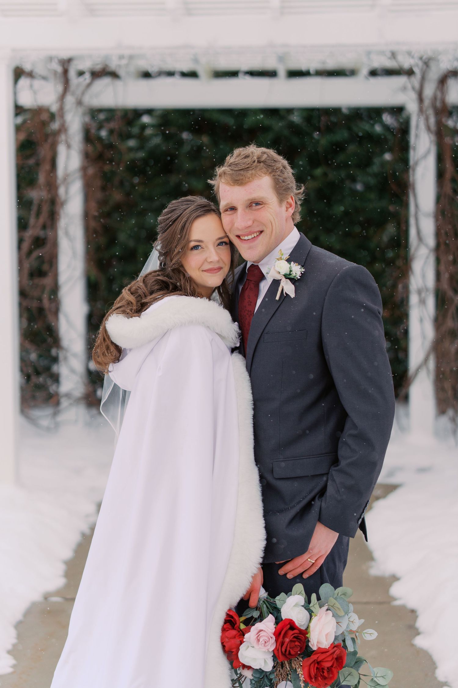 Newlyweds pose together under white arbor in snow with winter wedding bouquet.