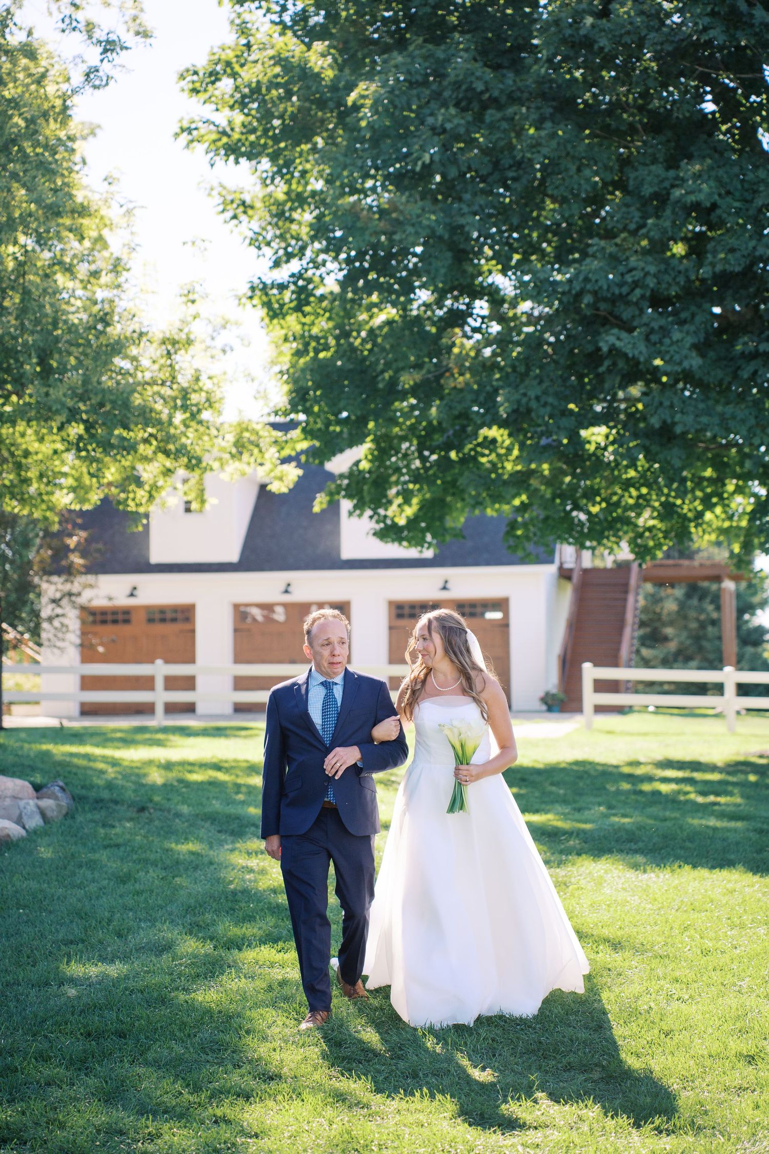 Wedding couple walks together on a sunny lawn with trees and a white building in the background.