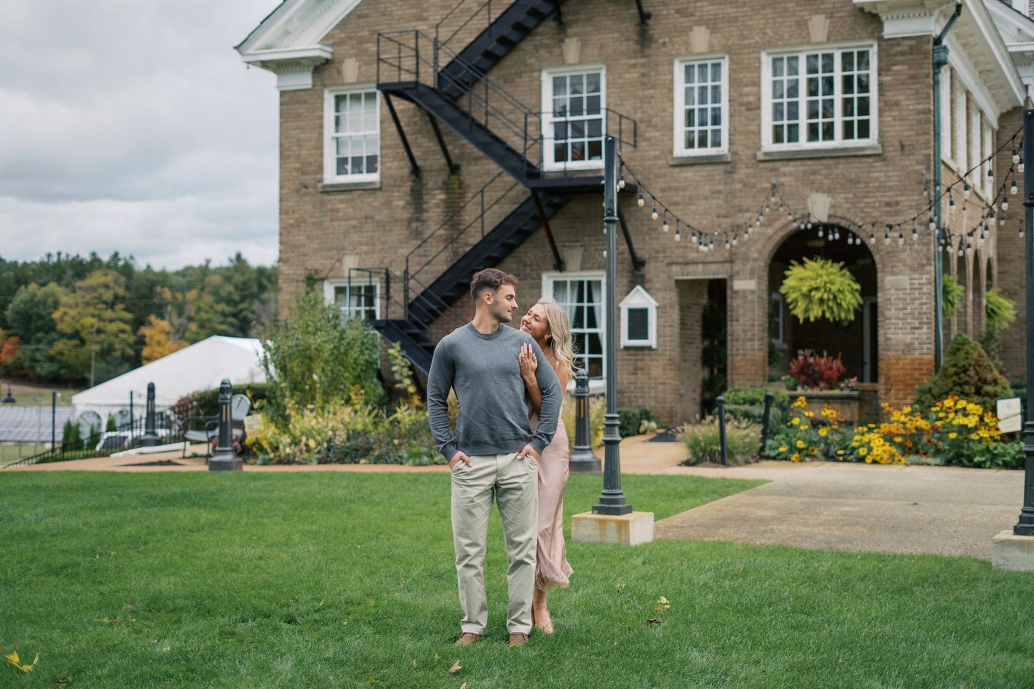 A couple stands together on a landscaped lawn outside a brick building with white trim windows.