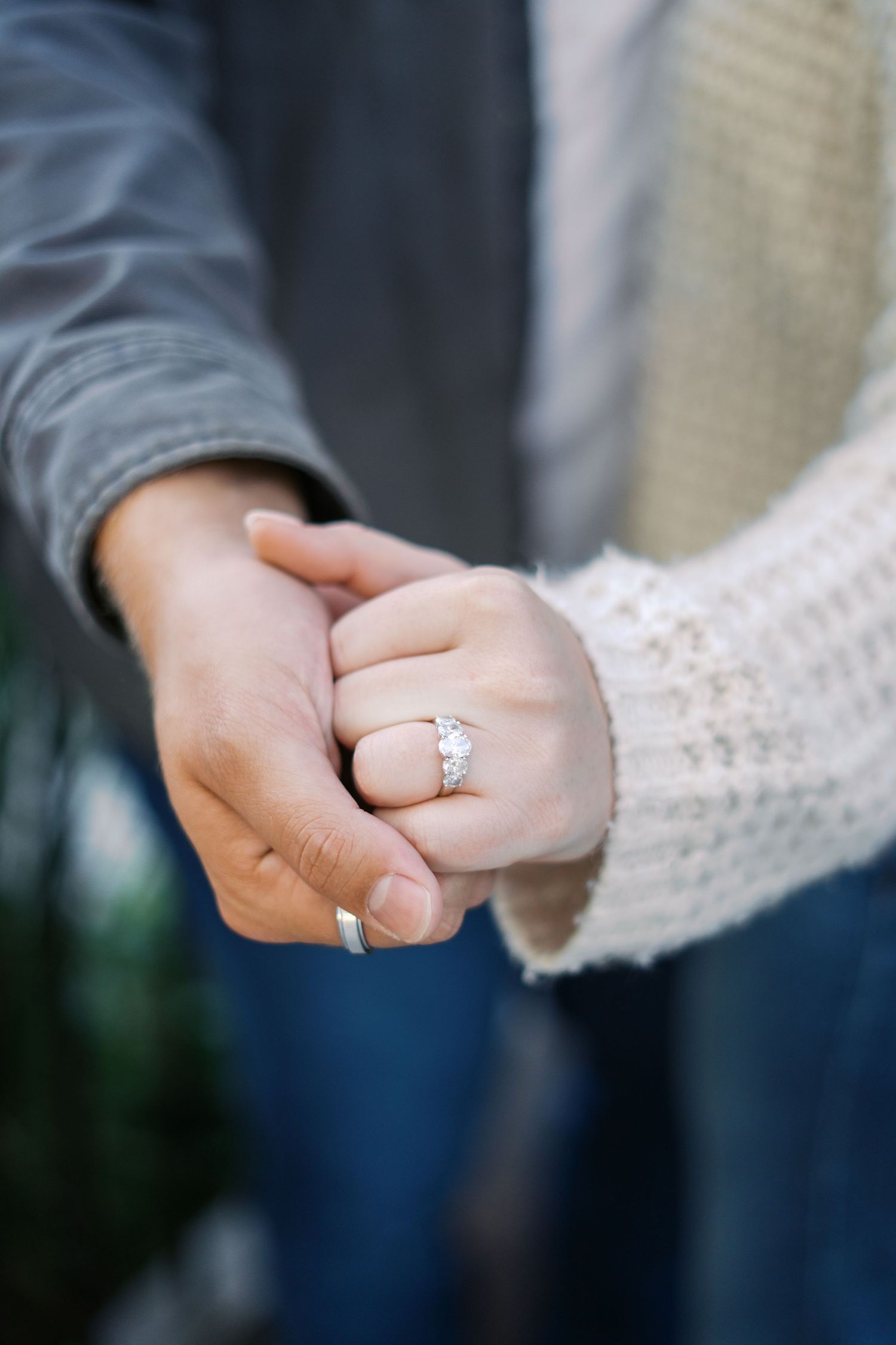 Close-up of hands showing off a sparkling engagement ring against a blurred background.