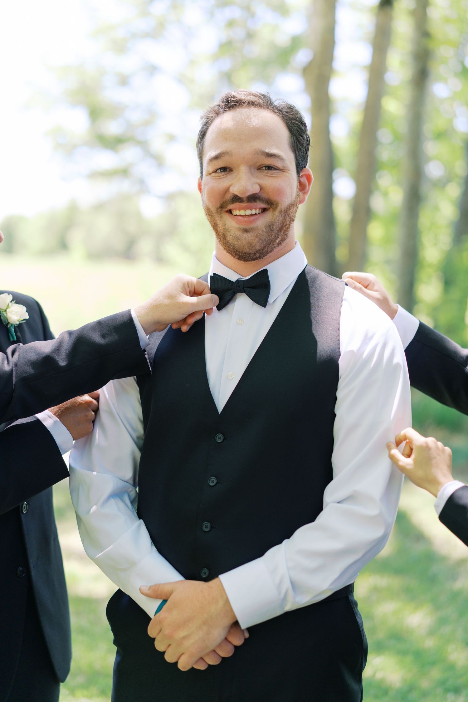 Smiling groom in black vest and bow tie being helped by groomsmen outdoors on wedding day.