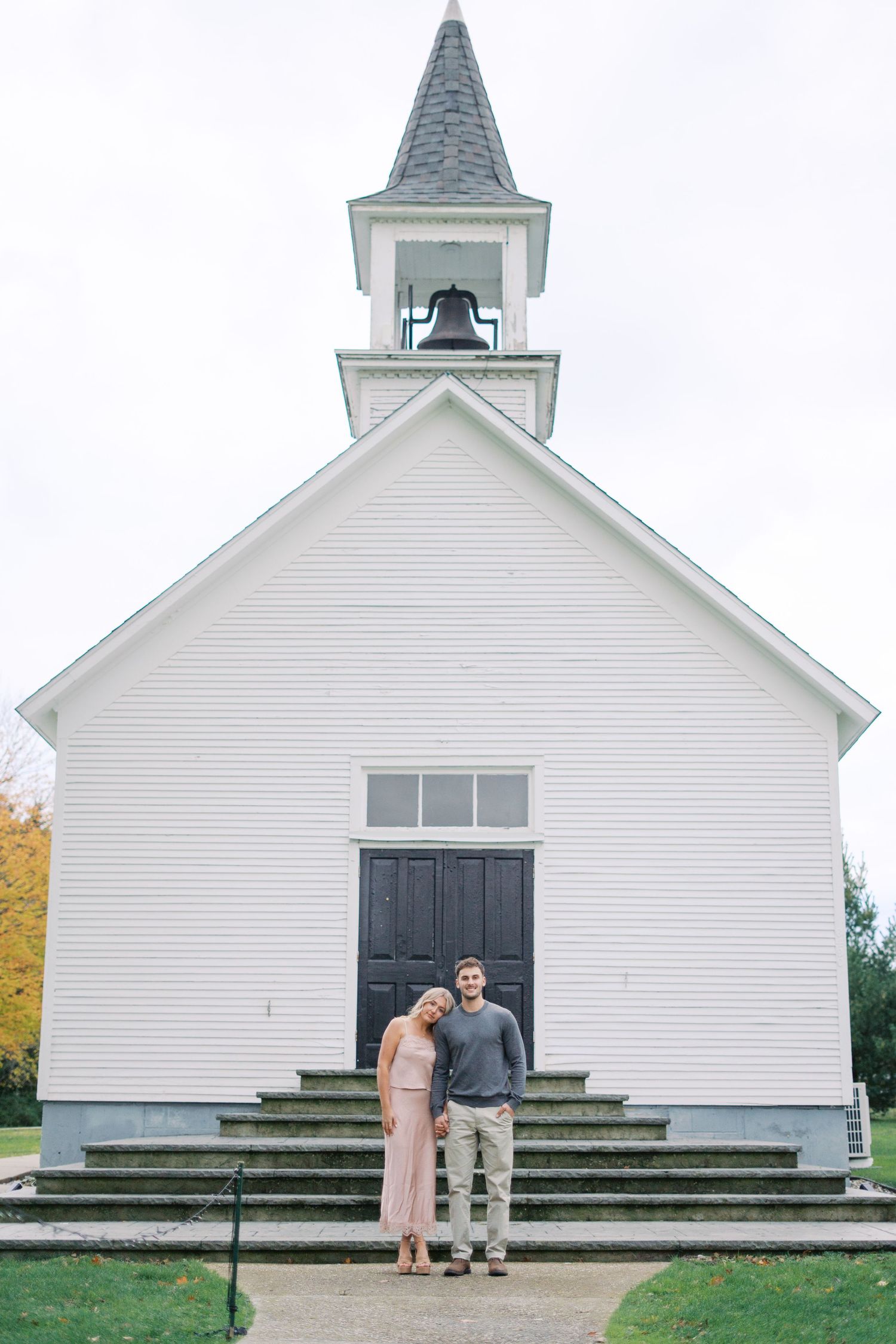 A couple stands together in front of a classic white church with a steeple.