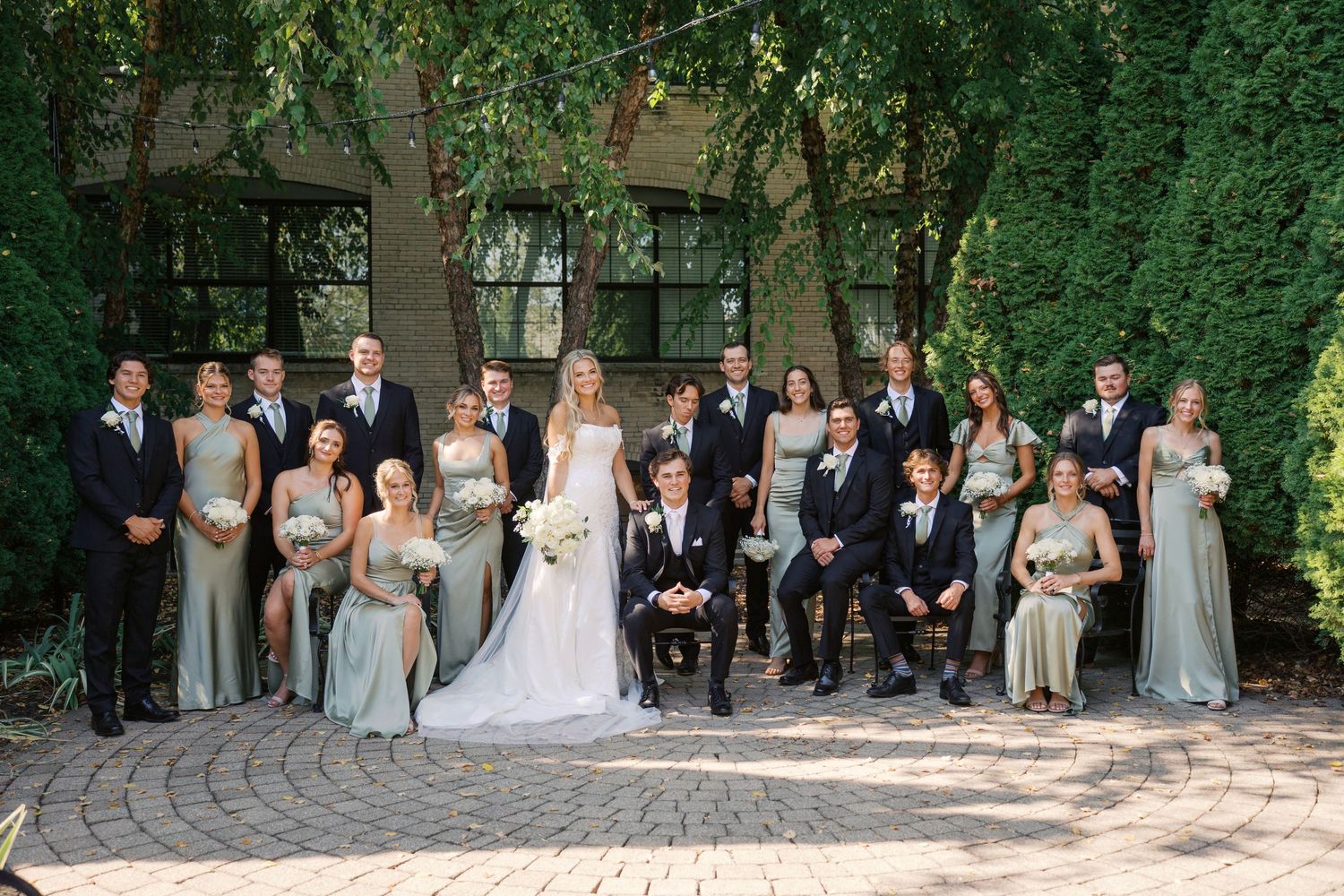 Full wedding party poses together on brick patio with green foliage backdrop.