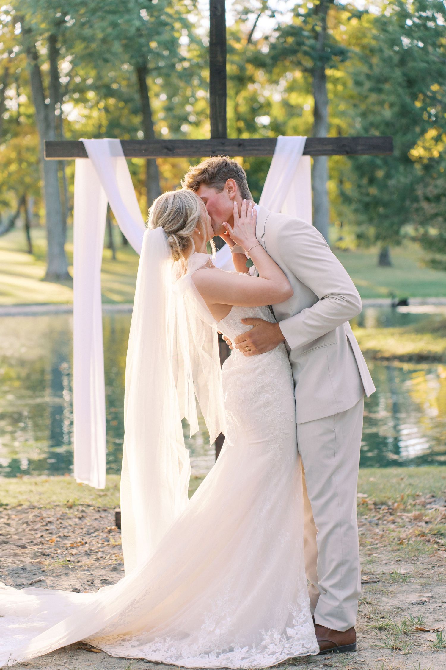 Romantic first kiss as newlyweds under wooden cross arch with flowing white fabric.