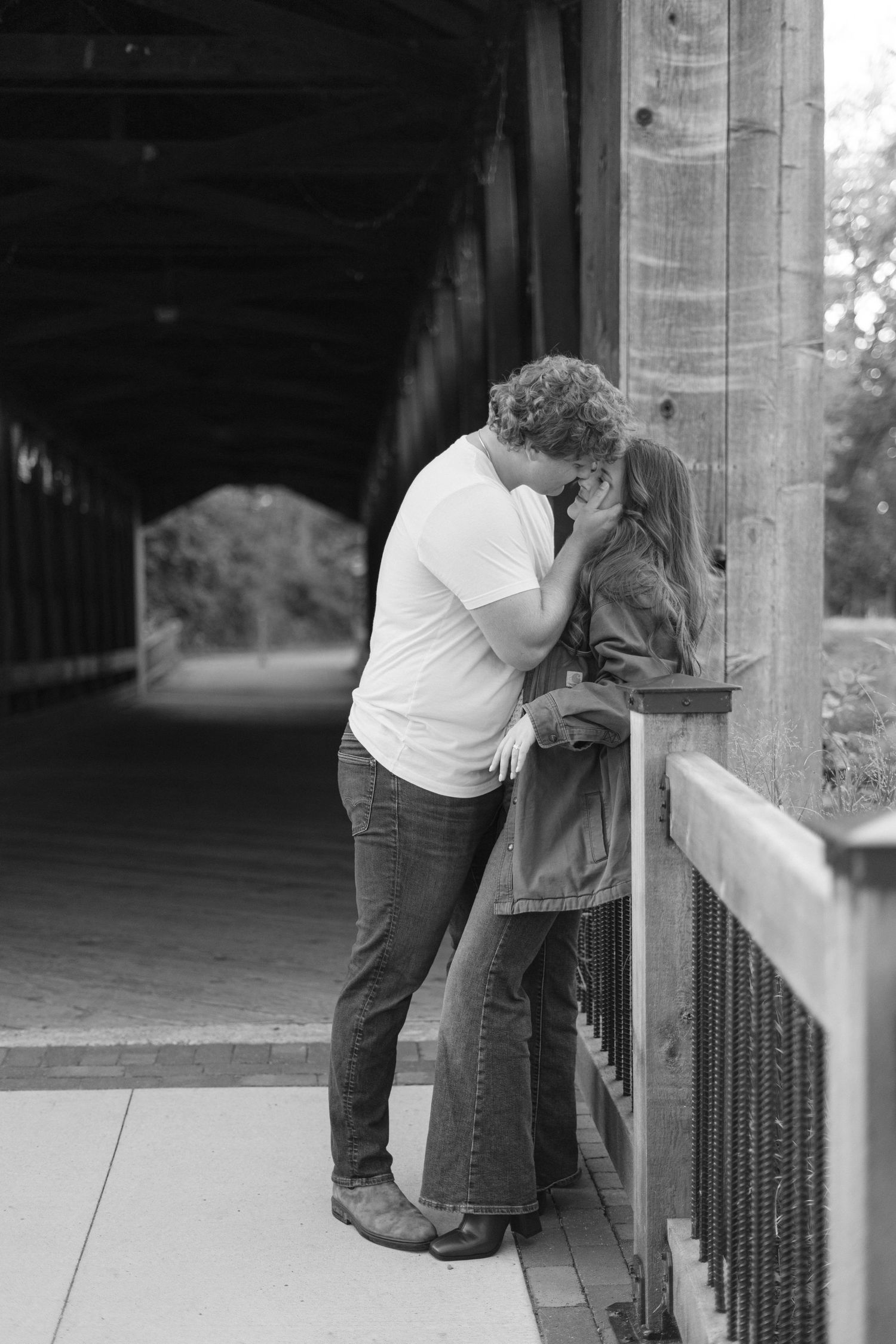 Couple leaning against wooden bridge post in black and white photograph.