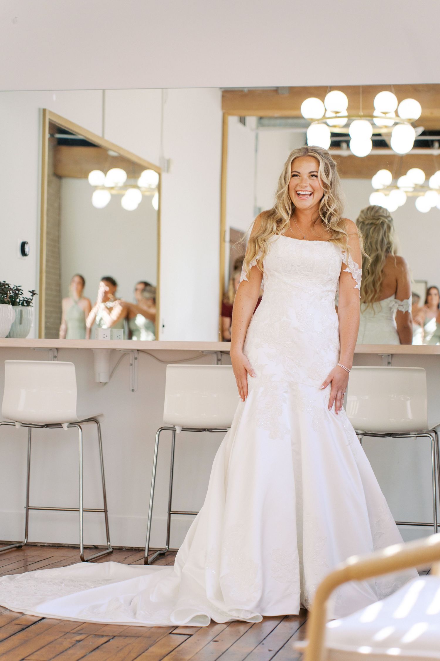 Bride in elegant white wedding gown poses in brightly lit dressing room.
