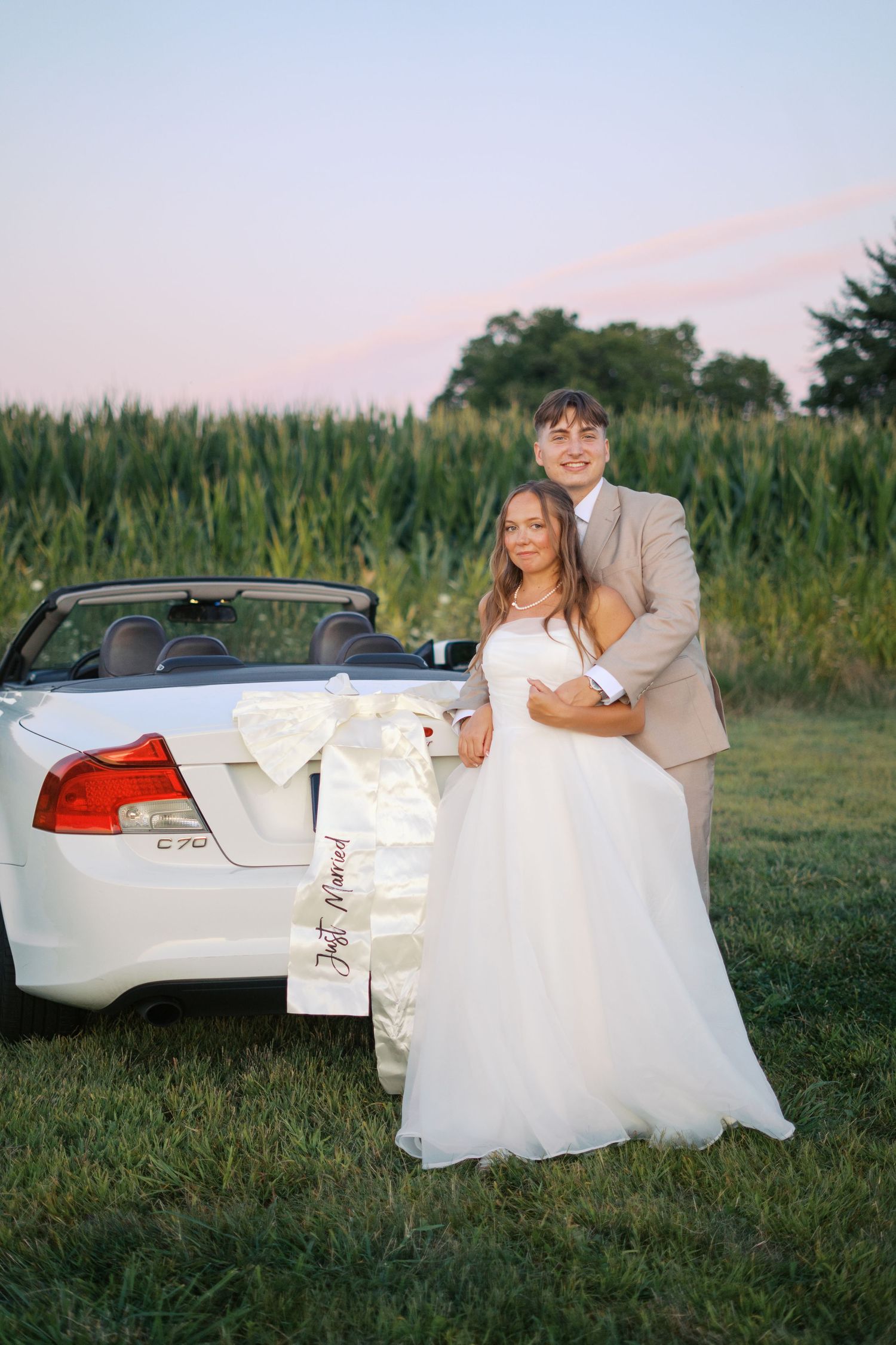 Bride in white dress and groom in tan suit pose playfully by their car in a rural setting.