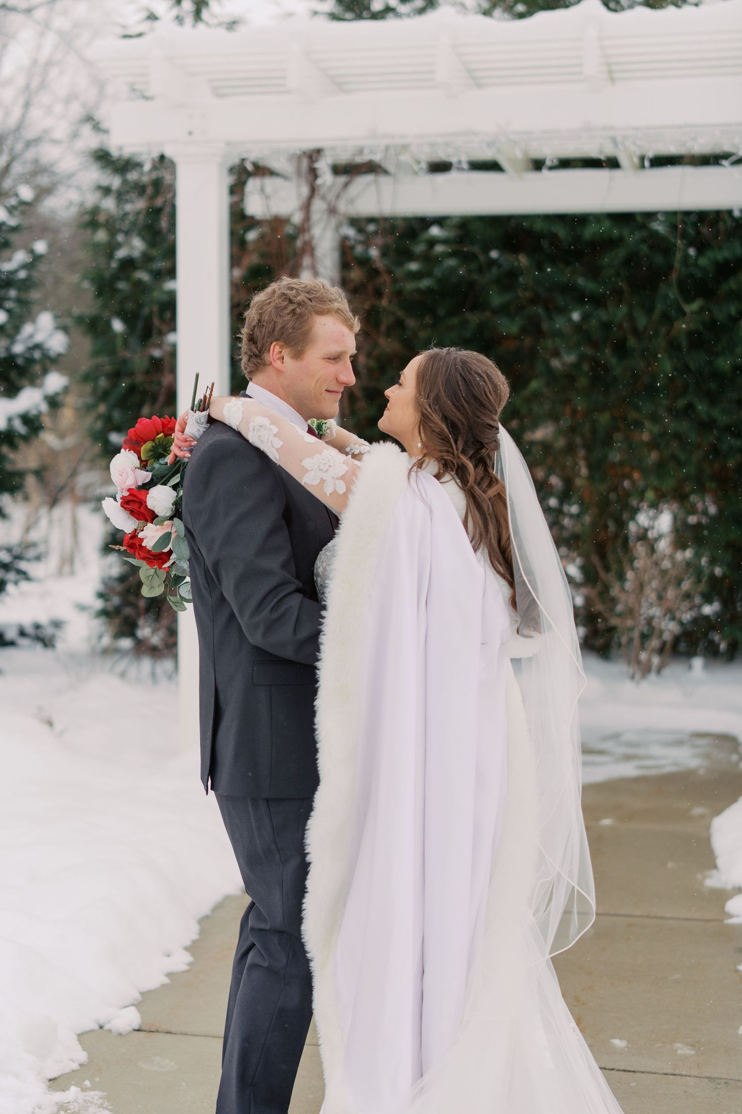 Winter wedding couple embracing under white pergola with snow backdrop.