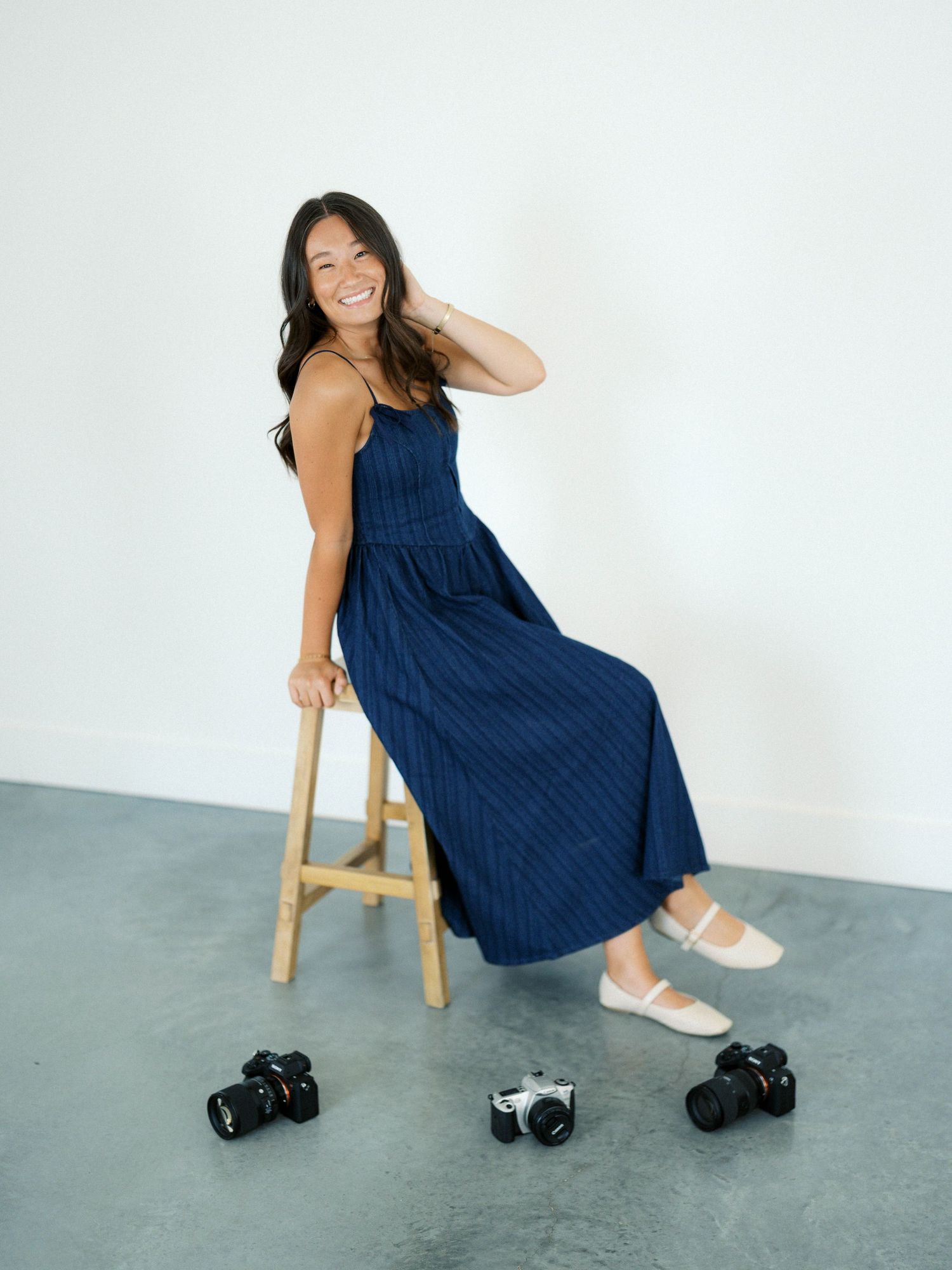 Smiling woman in navy striped maxi dress seated on wooden stool surrounded by cameras on white background.
