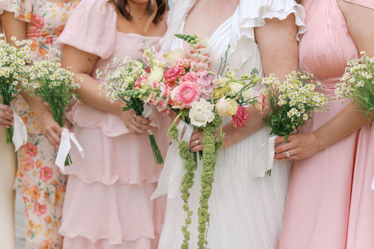 Bride and bridesmaids in pink dresses holding bouquets of pink roses and white wildflowers at a wedding ceremony.