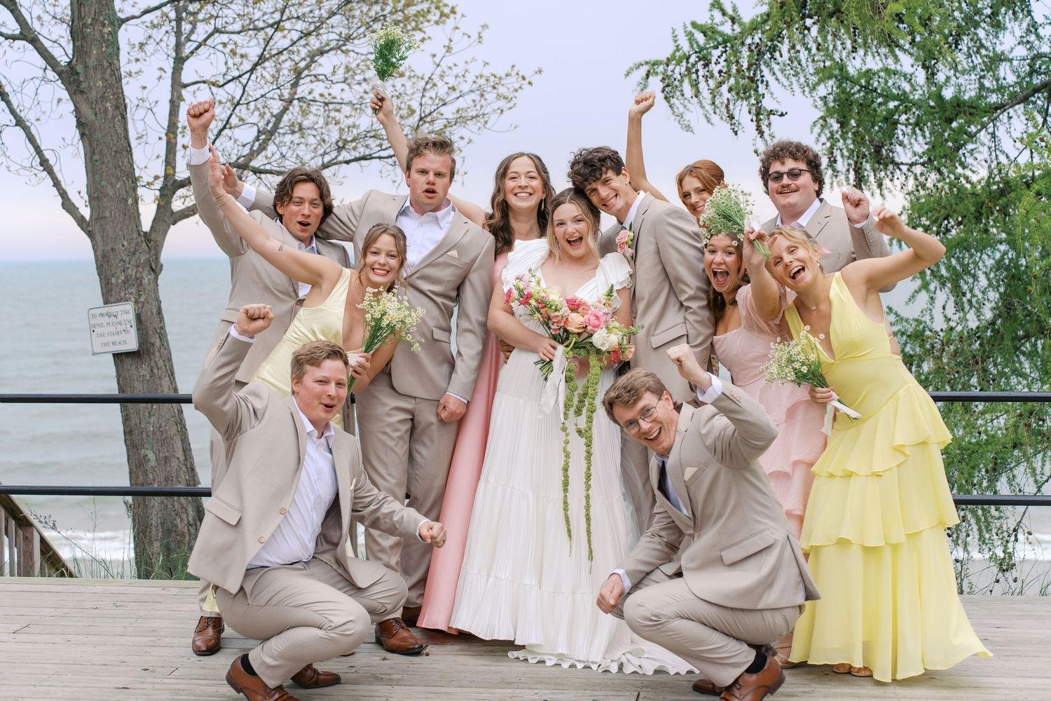 Joyful wedding party celebrating outdoors on a deck with ocean view, bride in white, bridesmaids in pink and yellow.
