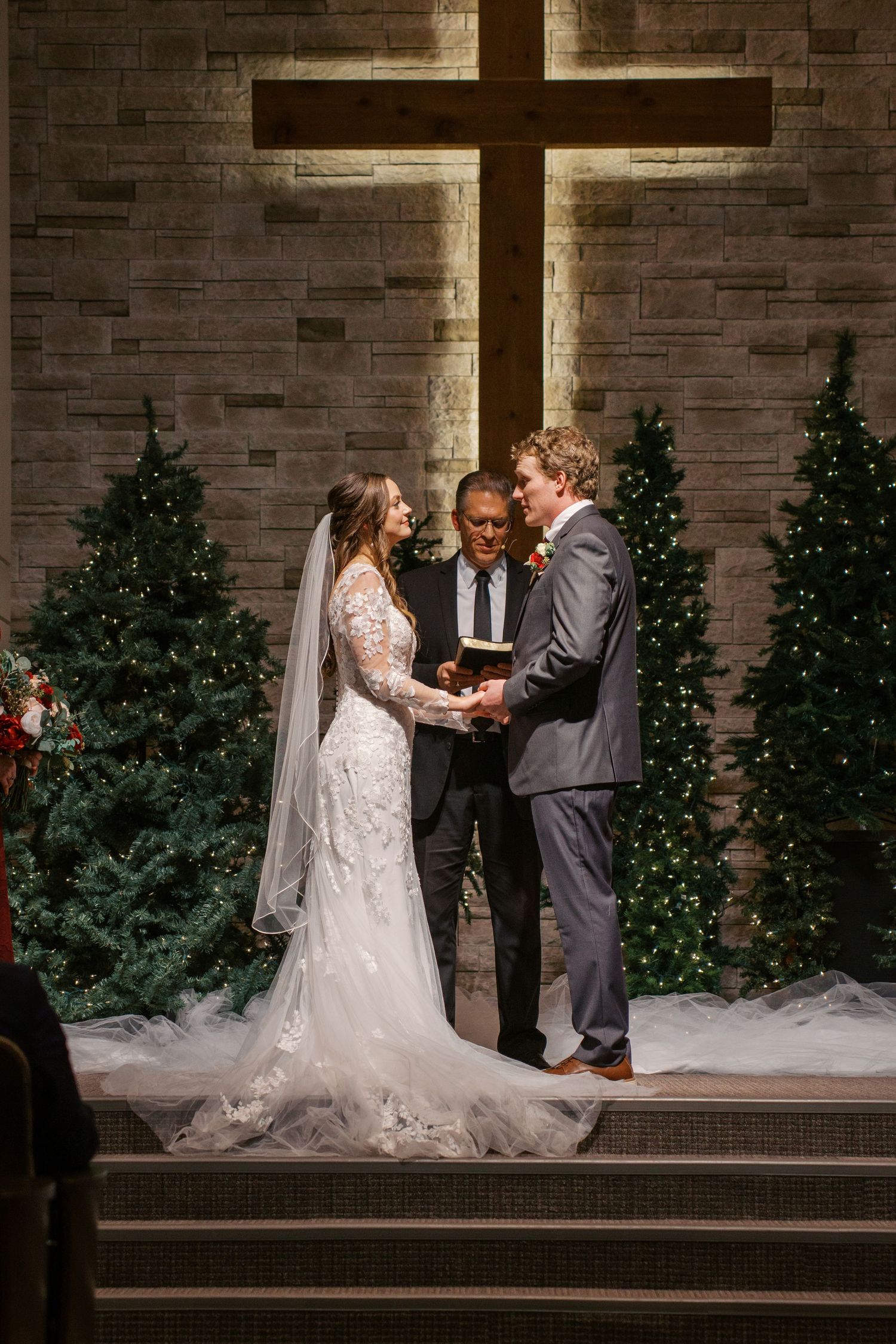 A wedding ceremony takes place in front of a brick wall with illuminated Christmas trees and a large cross.