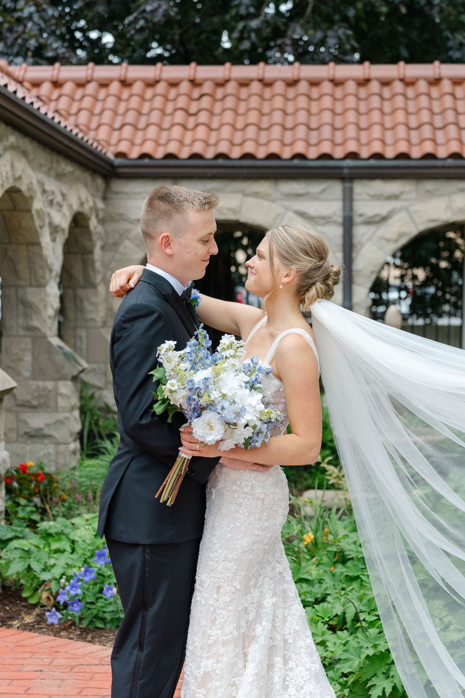 Romantic wedding portraits with flowing veil and blue floral bouquet against brick and stone architecture.