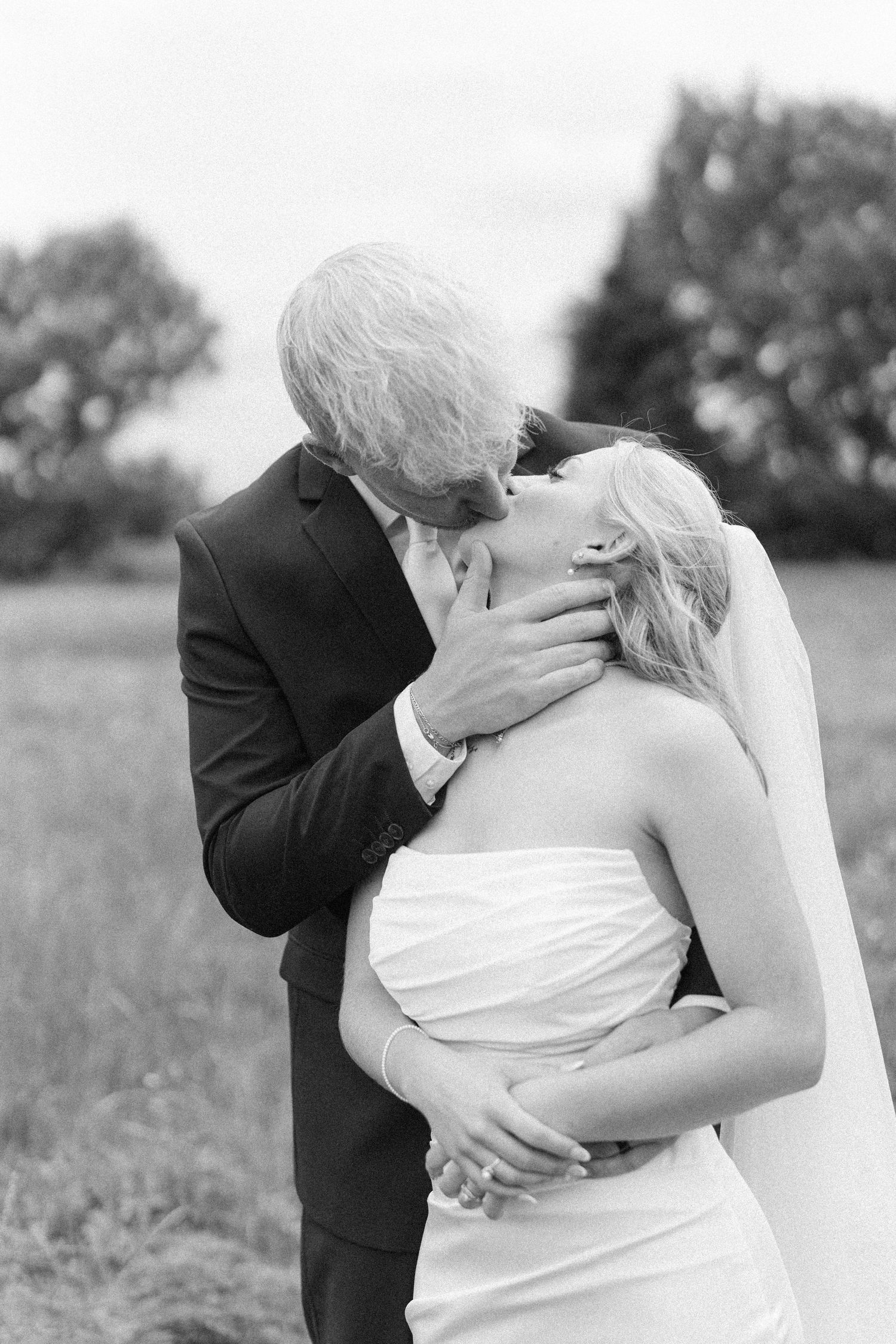 Black and white photo of a groom kissing his bride outdoors, with trees in the background.