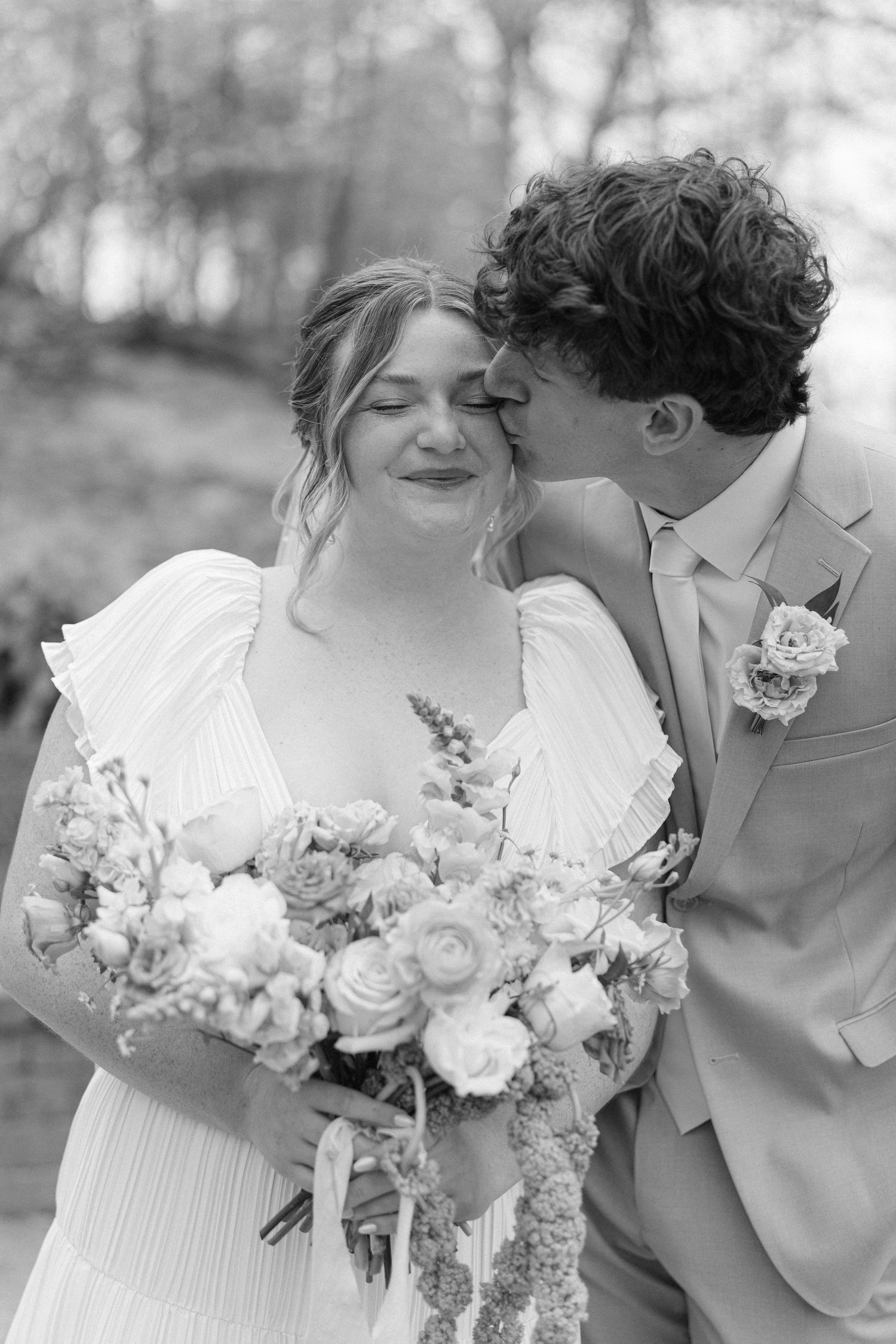 Black and white wedding photo of bride holding bouquet while groom kisses her cheek outdoors.