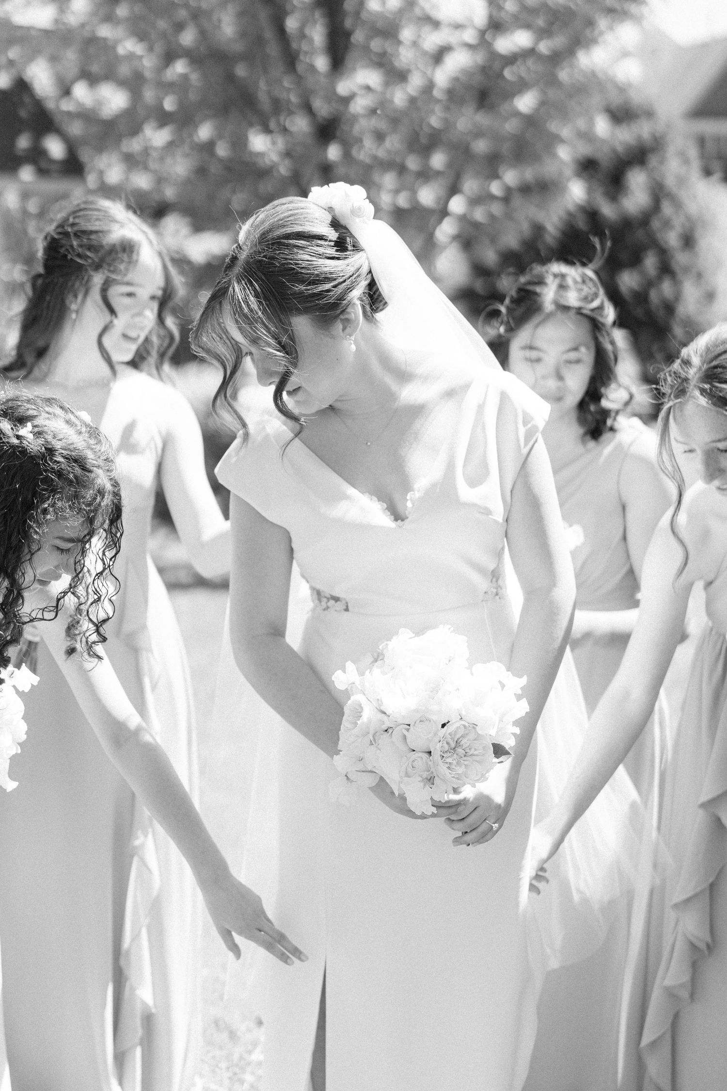Black and white photo of a bride holding a bouquet surrounded by smiling bridesmaids in flowing dresses outdoors.