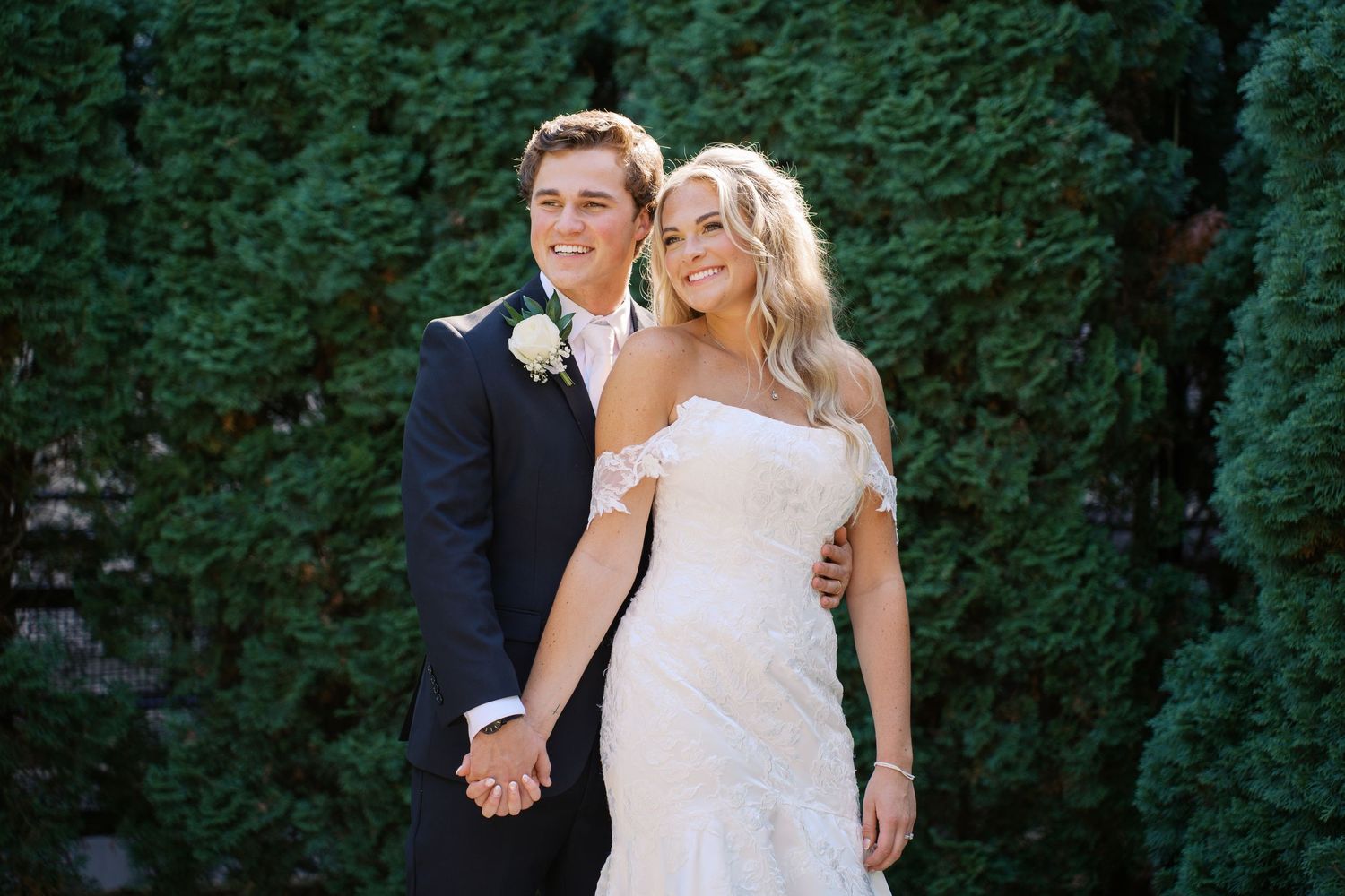 A couple in formal wedding attire poses together outdoors near green hedges.