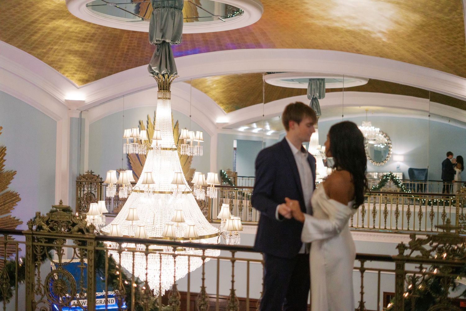 A couple dances together in an elegant ballroom with chandeliers and golden decor.