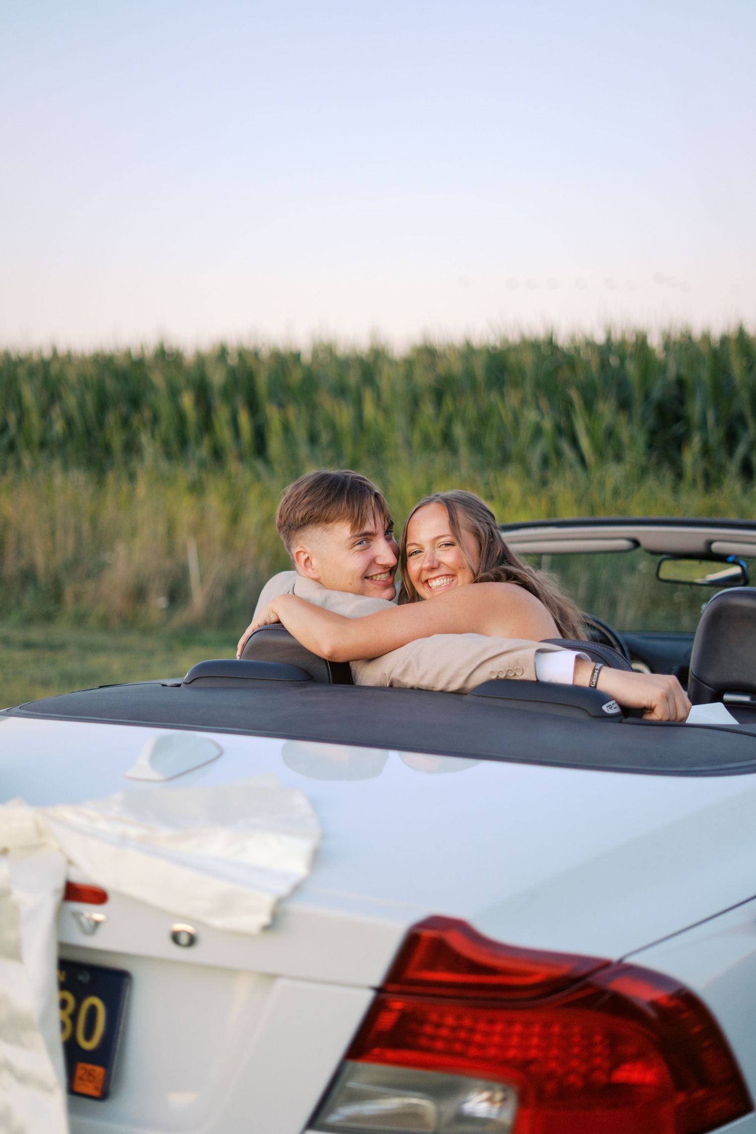 A couple shares an intimate moment while leaning against a white car at sunset with a cornfield in the background.