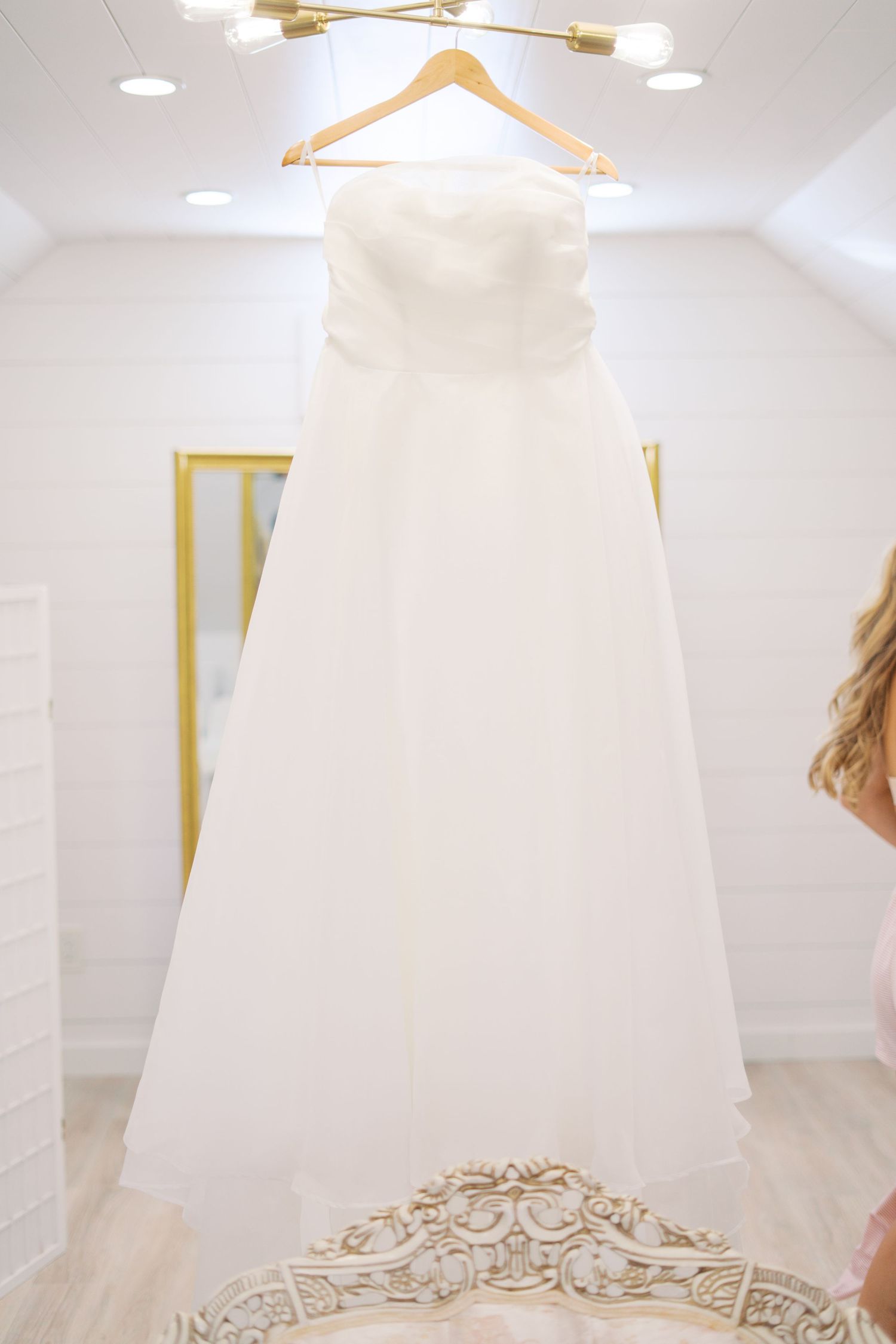 Elegant white wedding dress hanging in bright room with gold light fixtures.