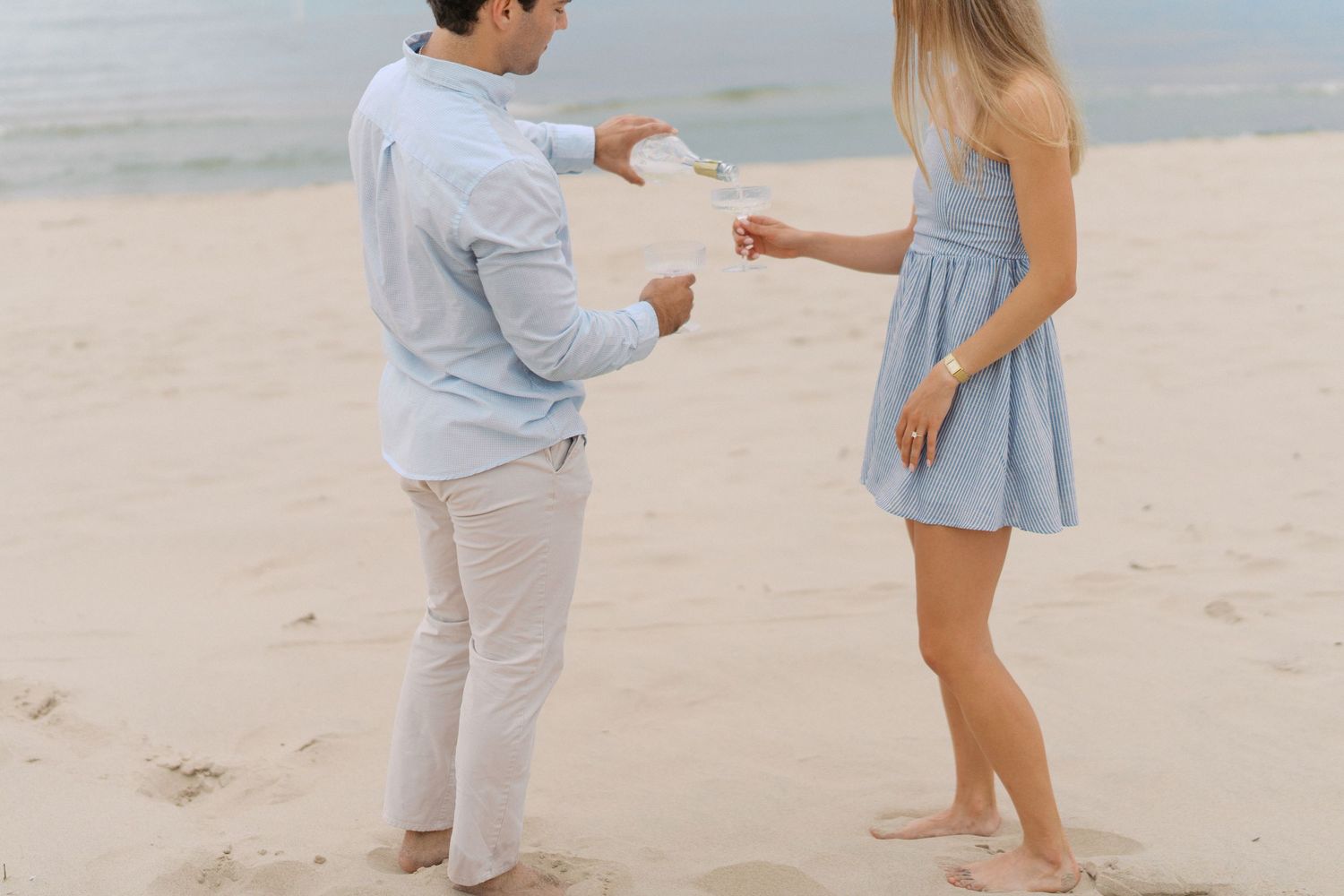 Couple sharing a romantic moment on a sandy beach, exchanging something small while standing barefoot near the ocean.