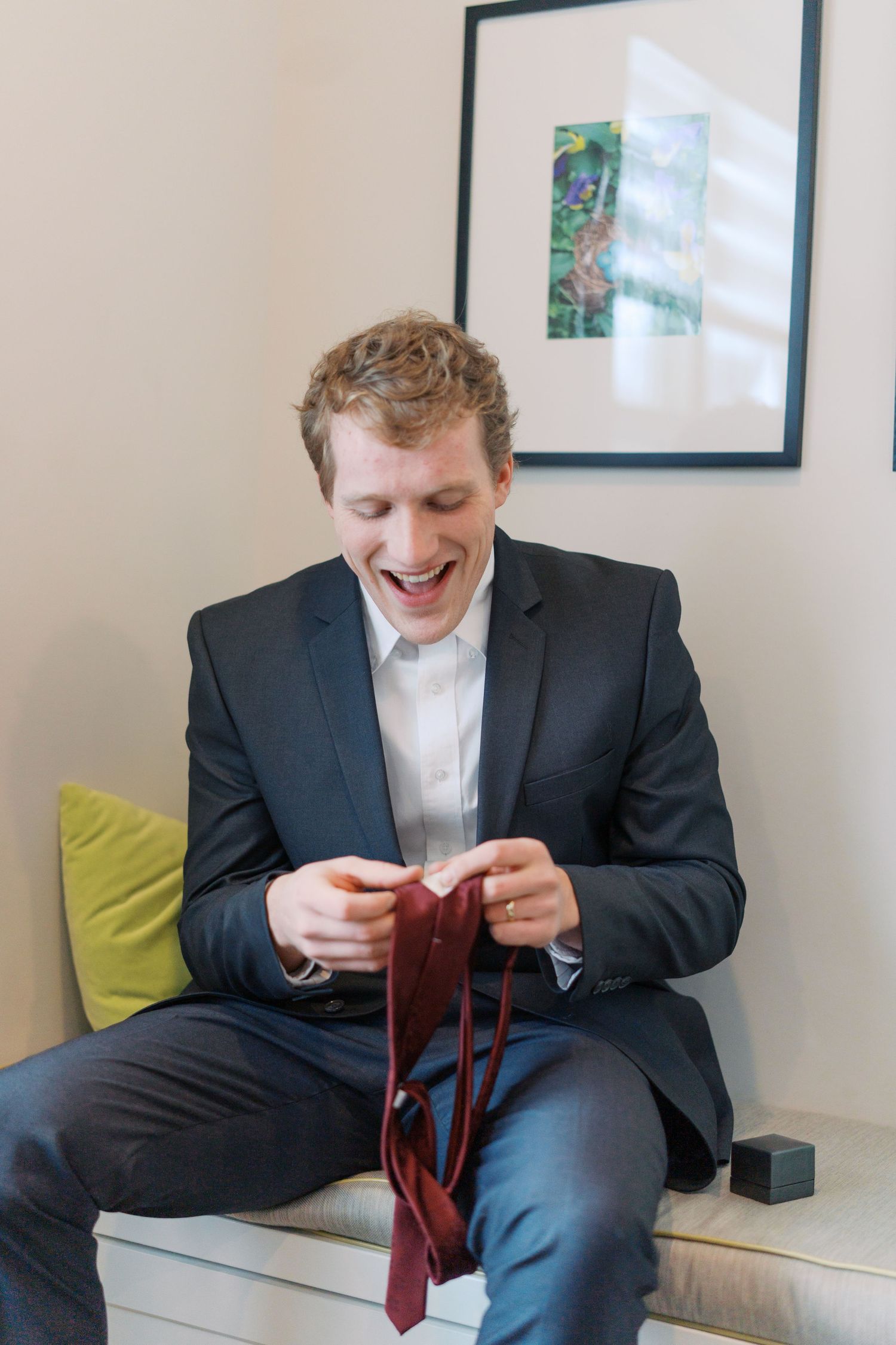 Groom adjusts his red tie while sitting on couch before wedding ceremony.