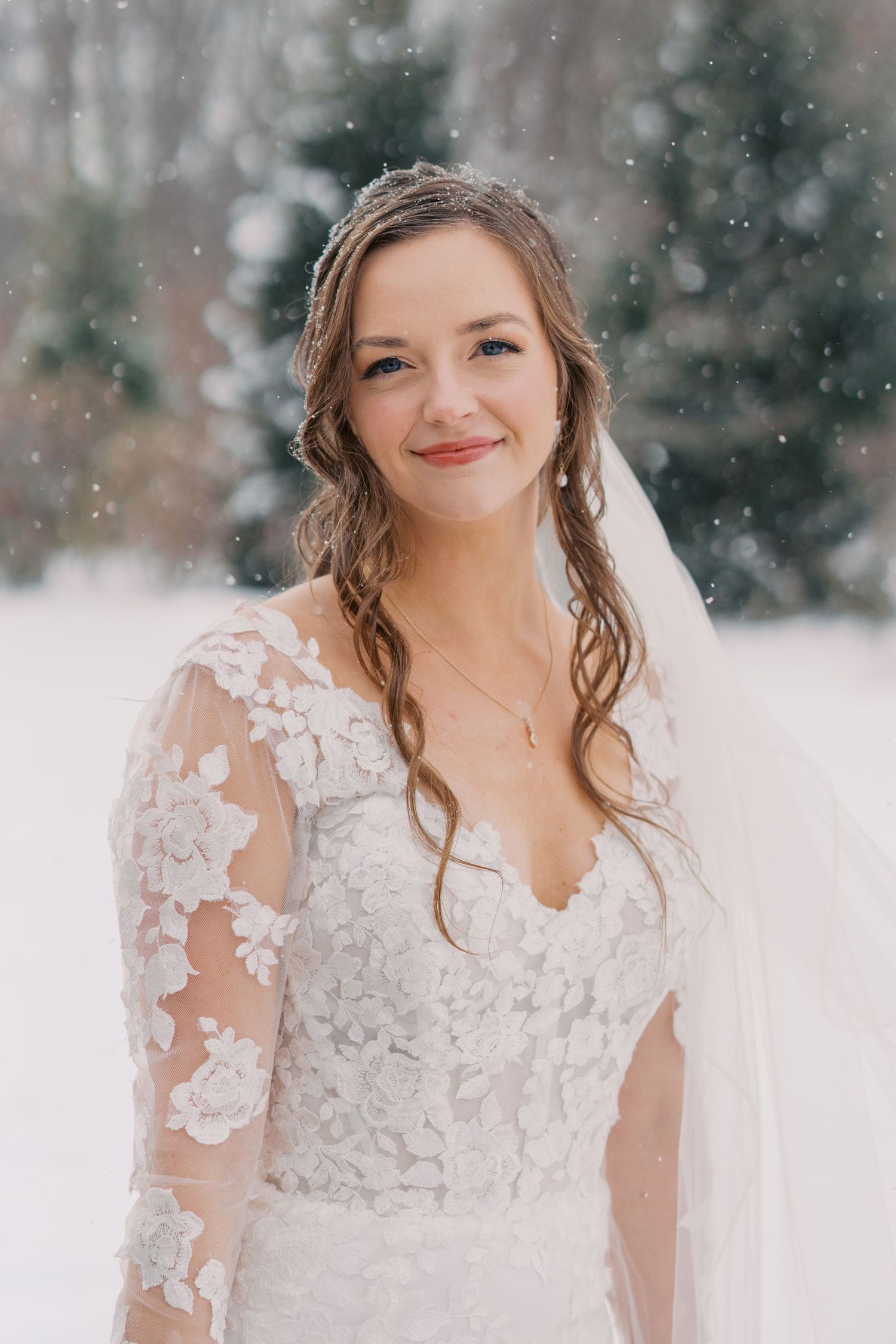 Bride in lace wedding dress poses in falling snow during winter wedding.