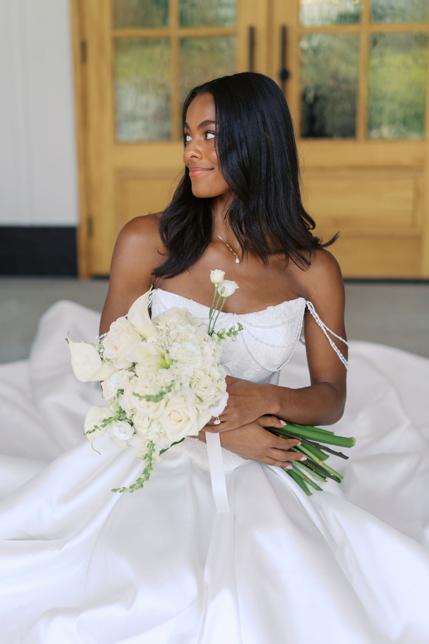Black bride in white off-shoulder ball gown holding white floral bouquet, gazing sideways near wooden doors.